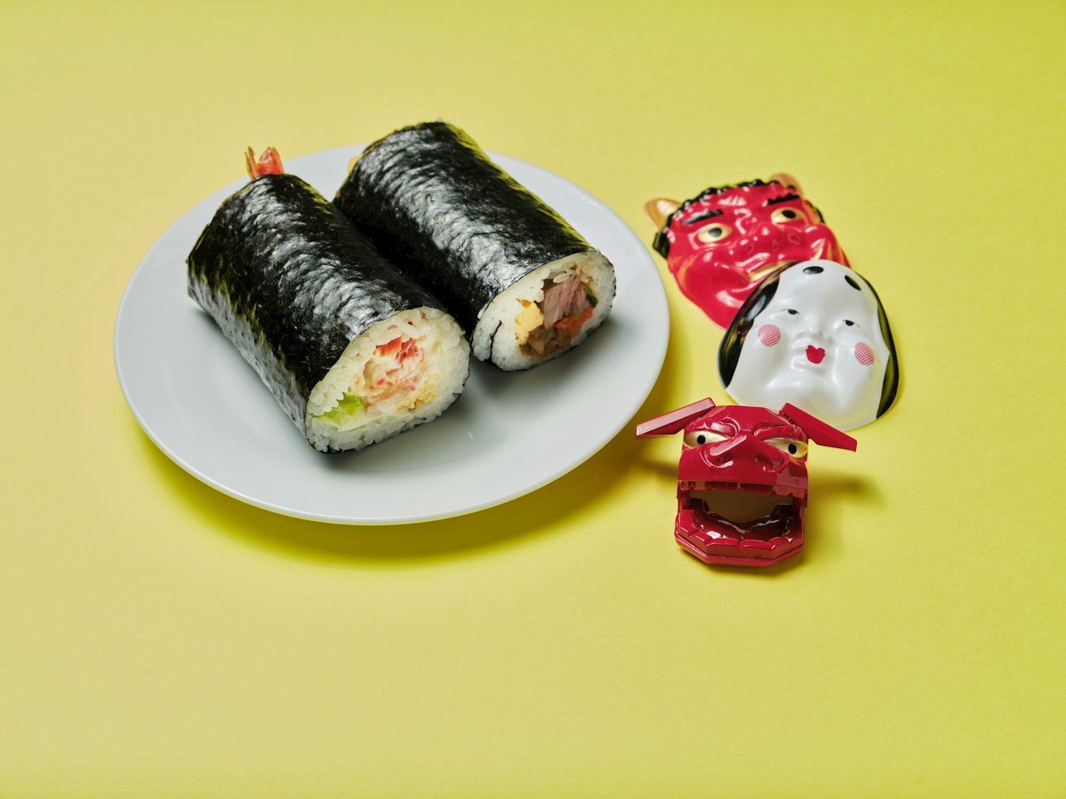 Gulung sushi di piring putih dengan figur dekoratif merah di latar belakang kuning