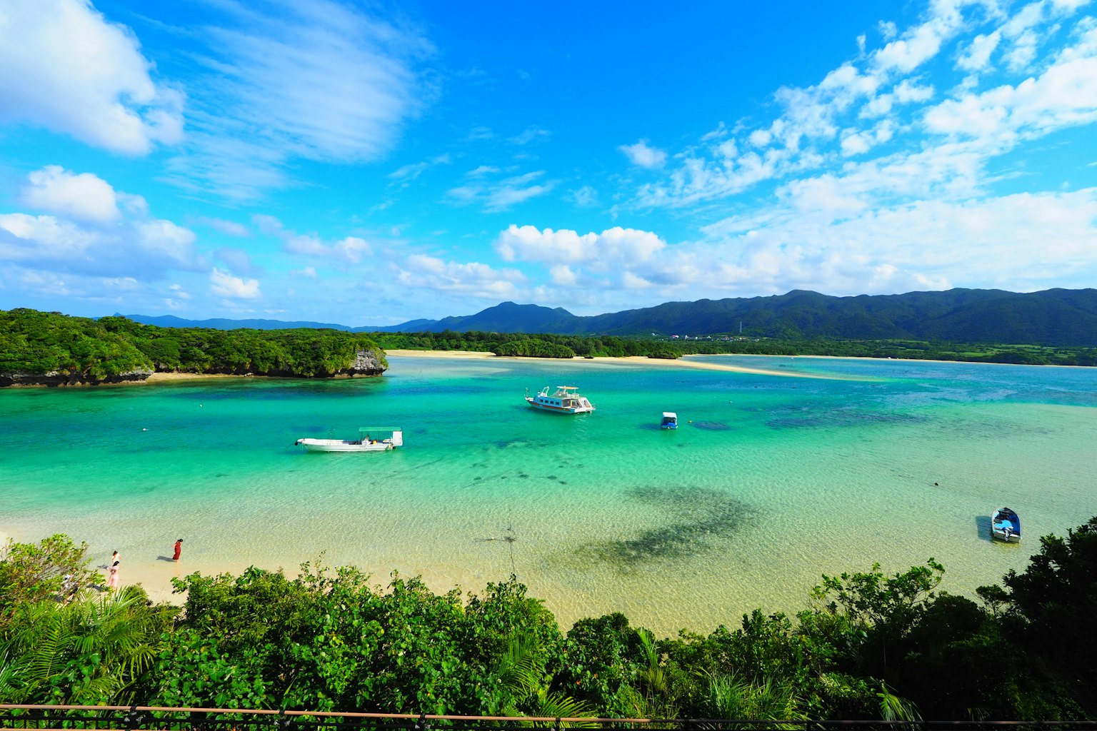 Vista panoramica di una baia turchese con barche e spiaggia di sabbia bianca circondata da vegetazione e cielo blu