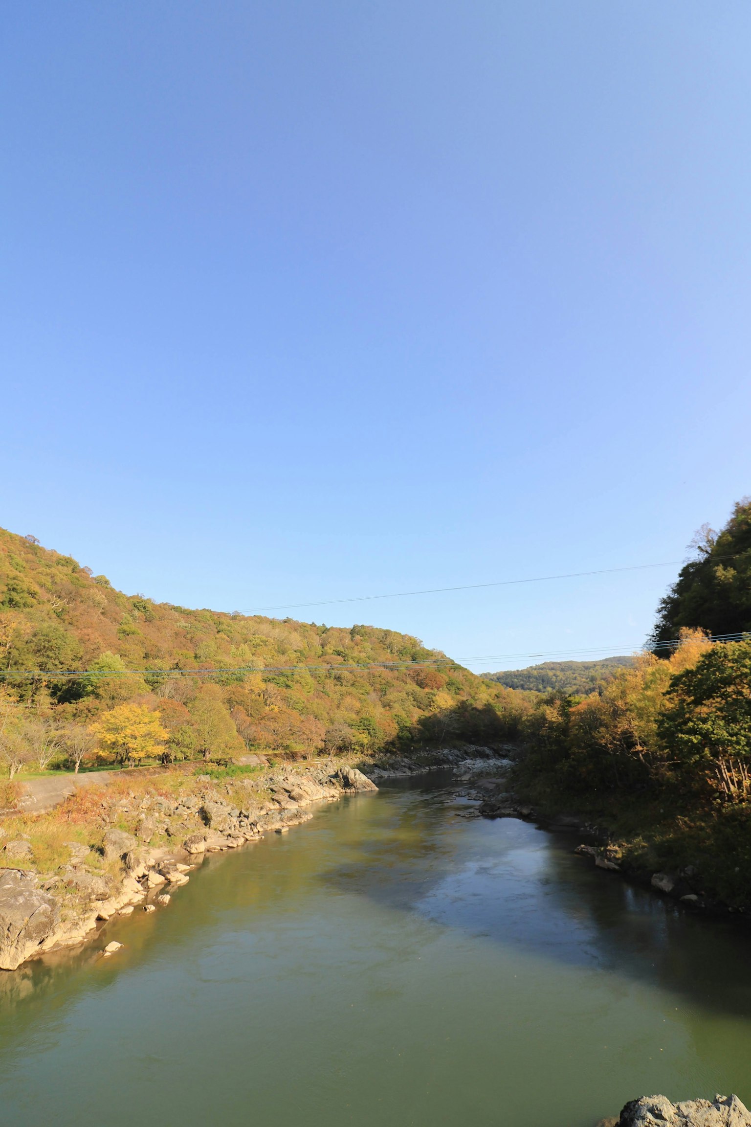 Paesaggio sereno di un fiume circondato da montagne dai colori autunnali sotto un cielo blu chiaro
