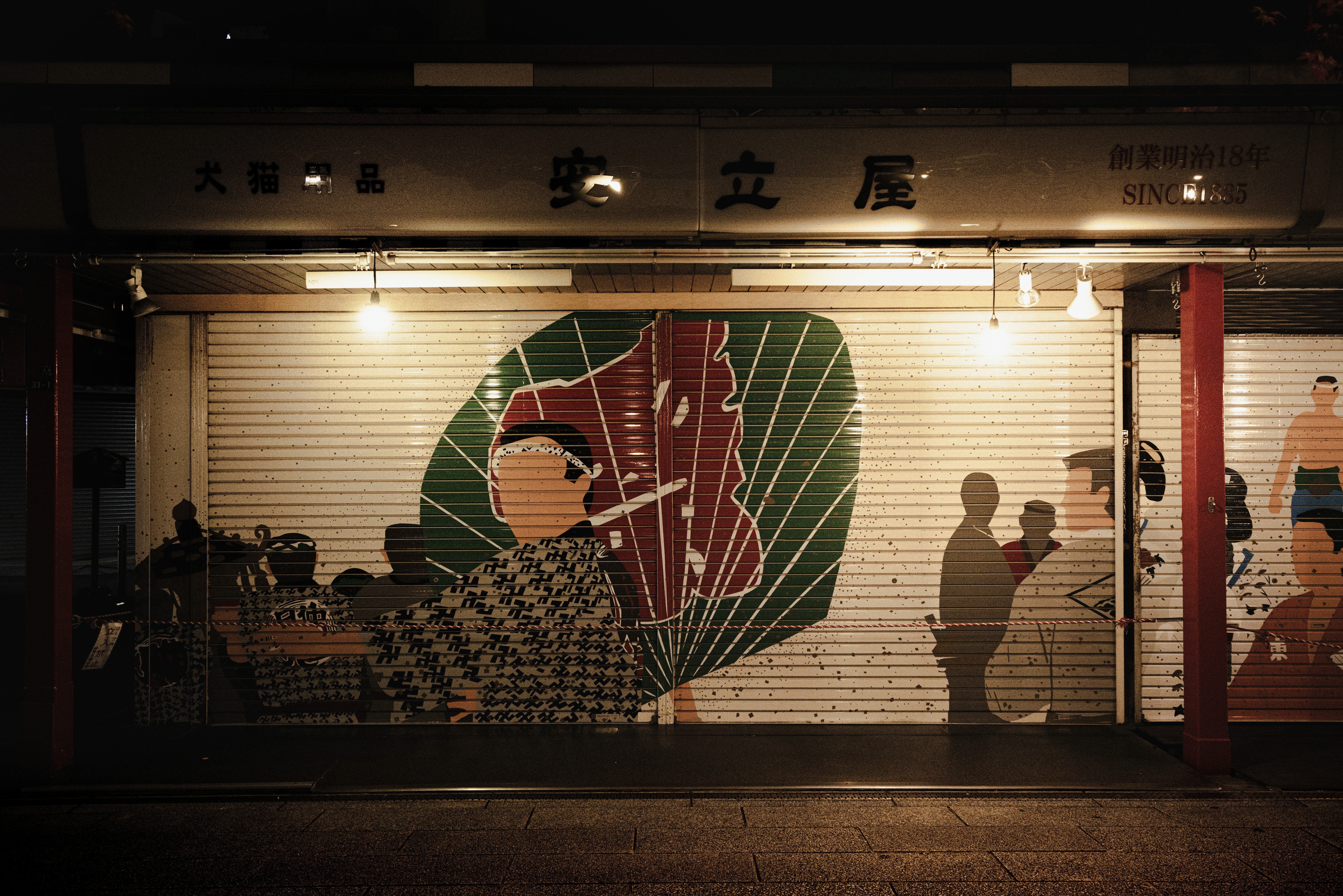 夜の街にある大きな扇子の絵が描かれたシャッターの前に人々が立っている