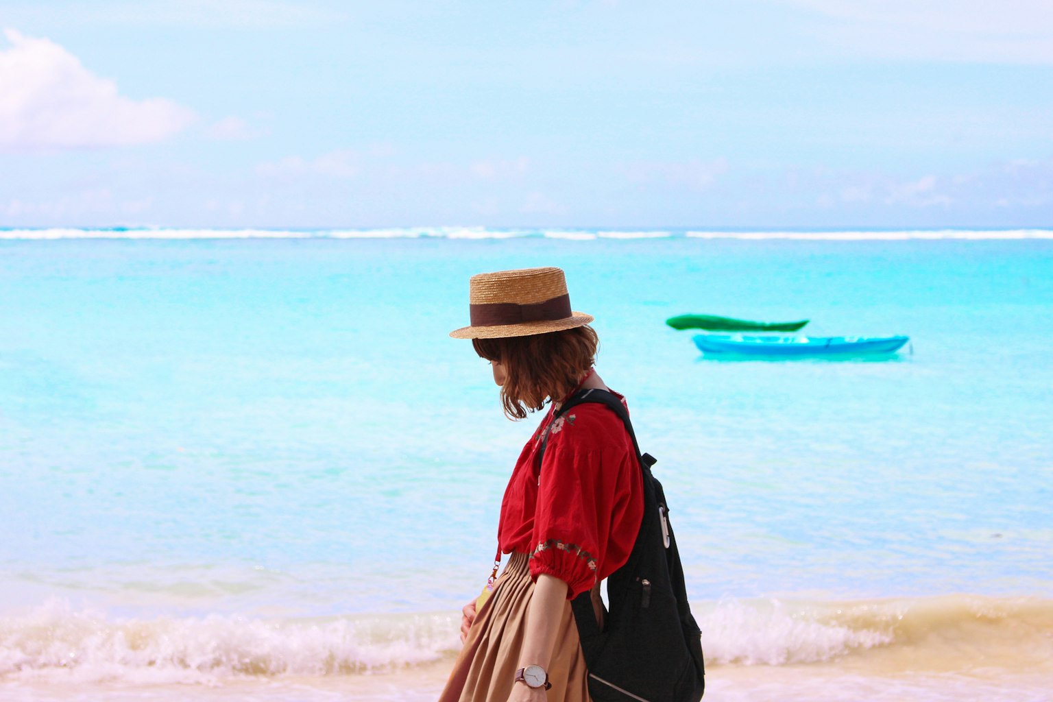 A woman wearing a hat walking along the beach with turquoise water and a kayak in the background