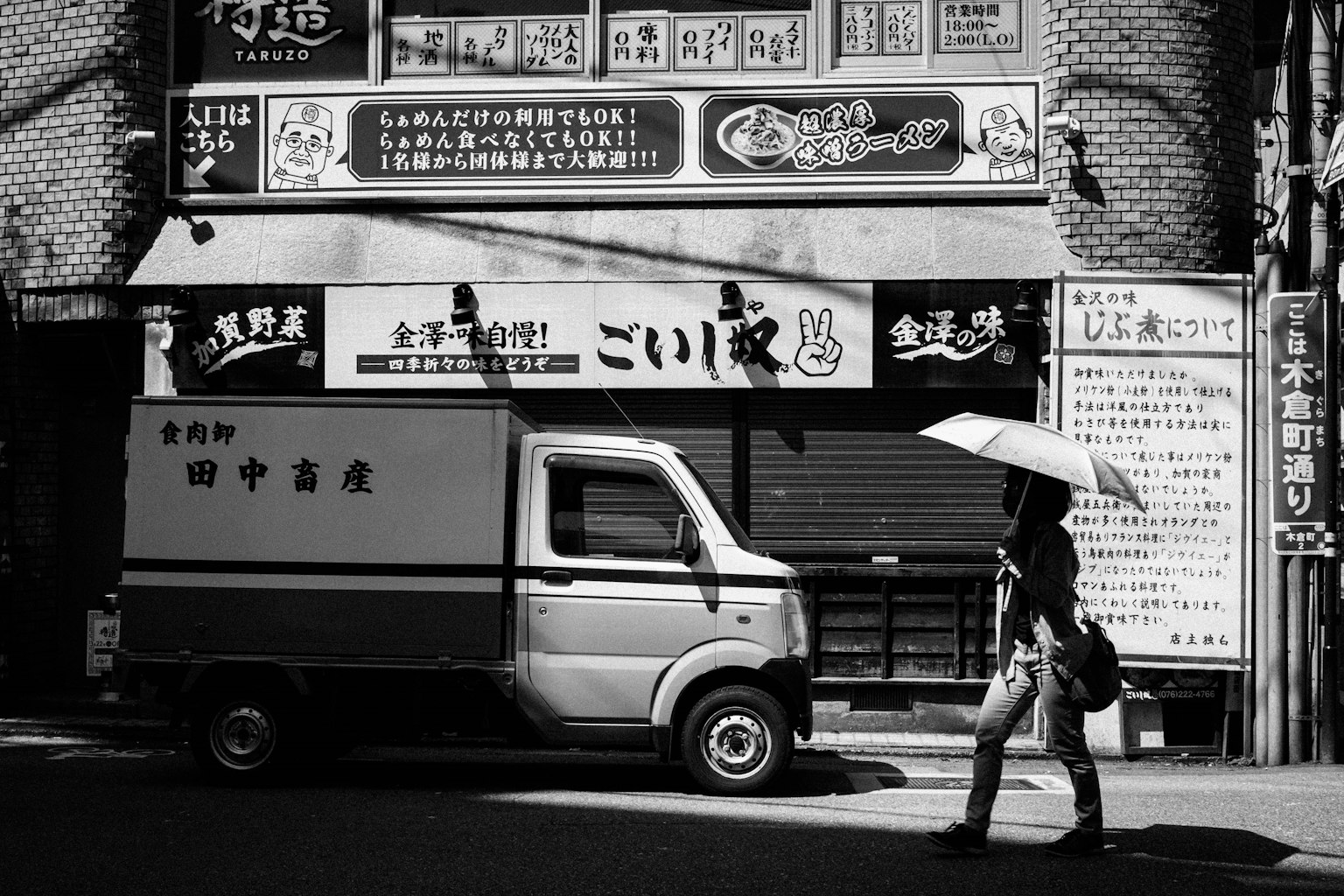 A black and white street scene featuring a person walking with an umbrella and a delivery truck in a commercial area