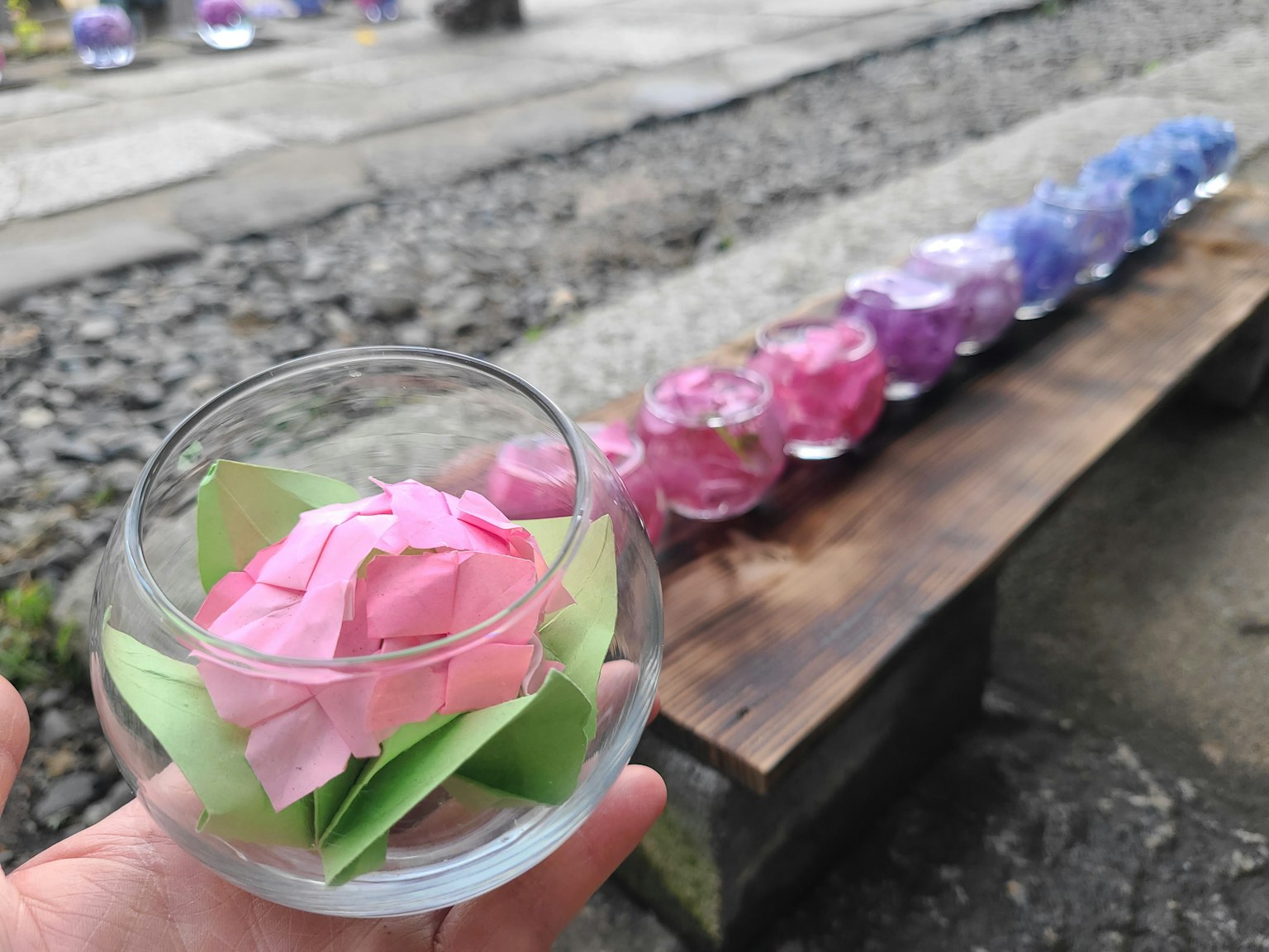 Una mano sosteniendo un tazón de vidrio transparente con una flor rosa y hojas verdes adentro mientras velas de colores están alineadas al fondo