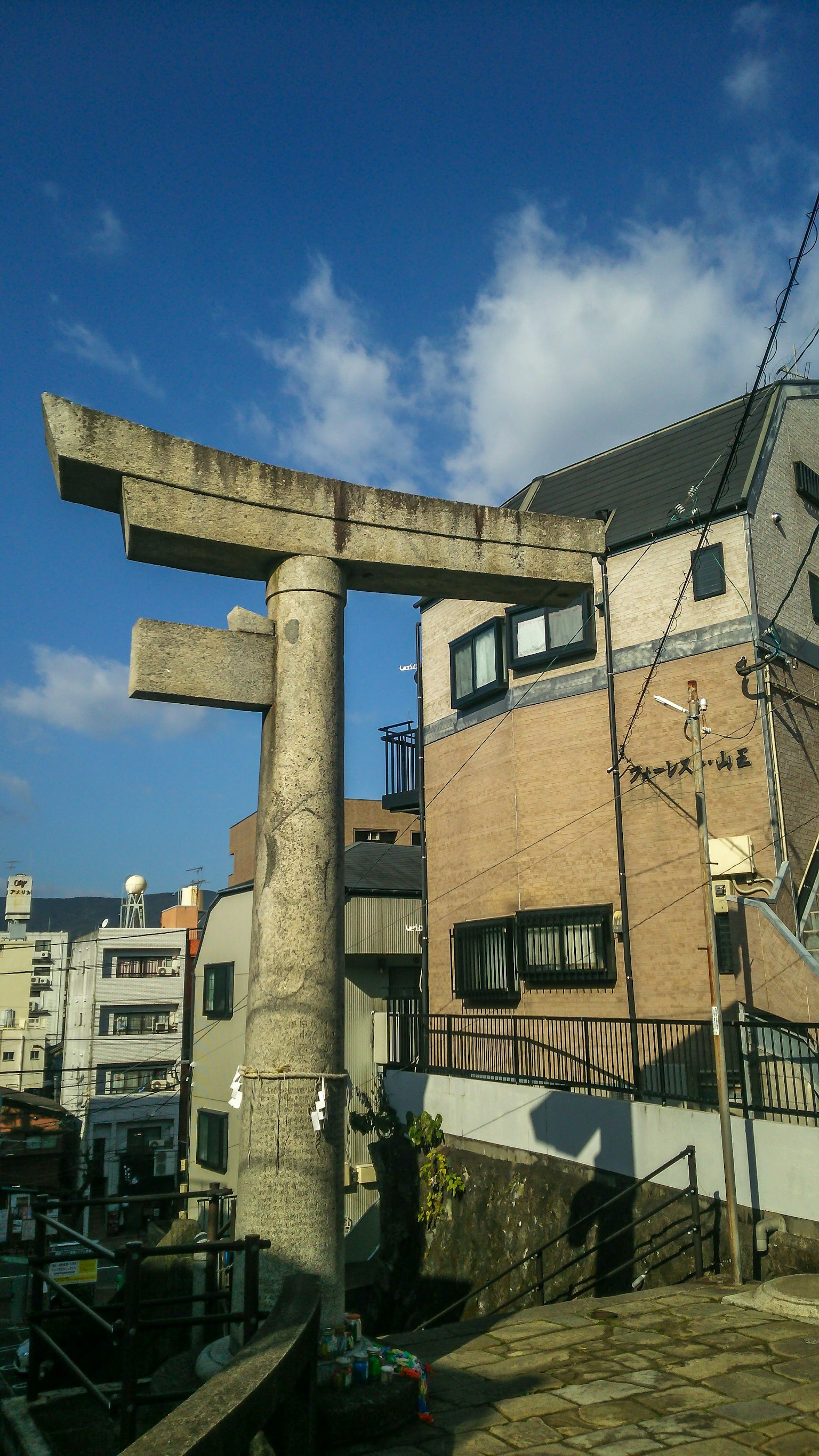 Cổng torii bằng bê tông với các tòa nhà xung quanh dưới bầu trời xanh