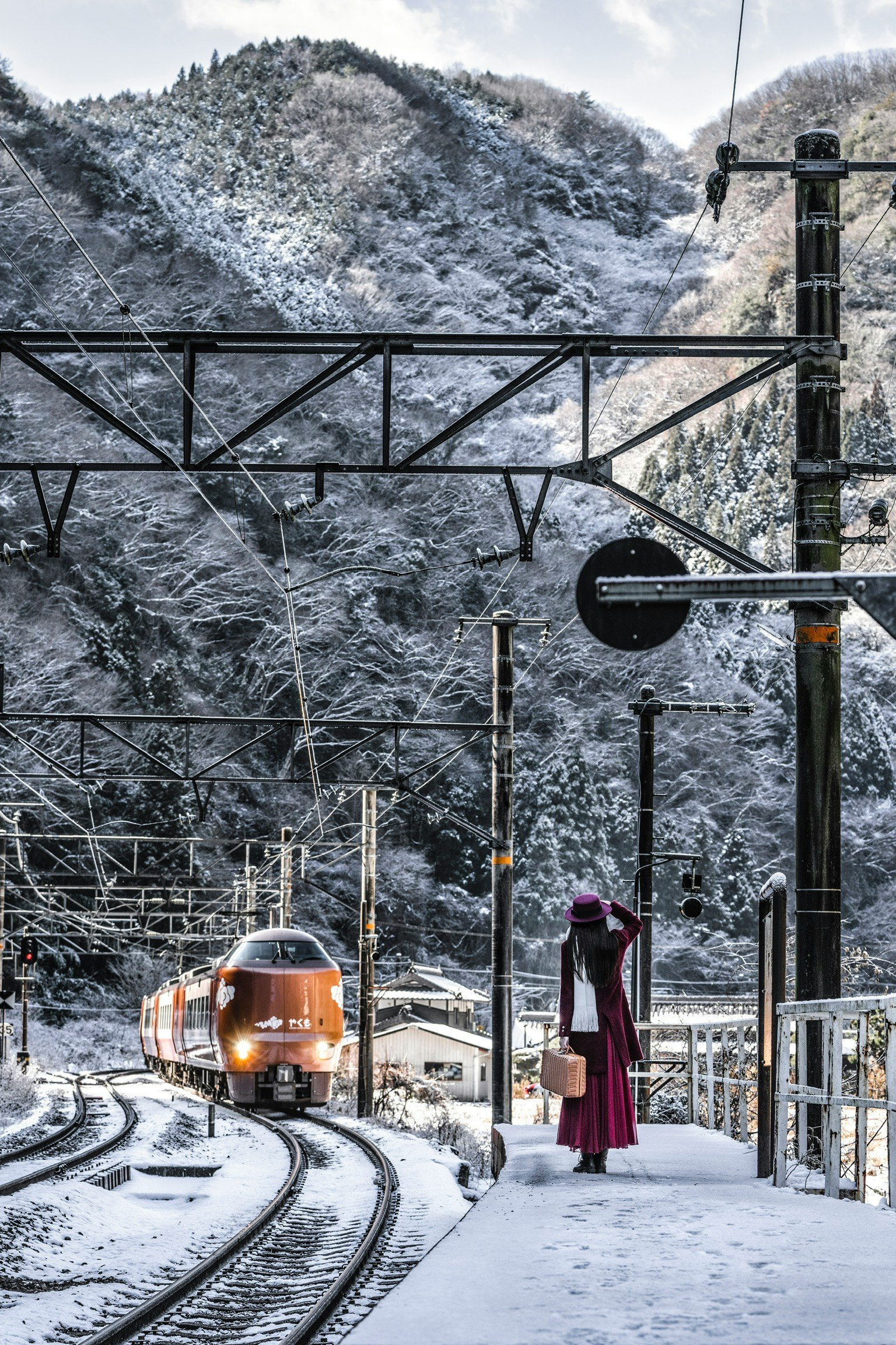 Tren llegando a una estación nevada con montañas al fondo y una persona vestida de rojo