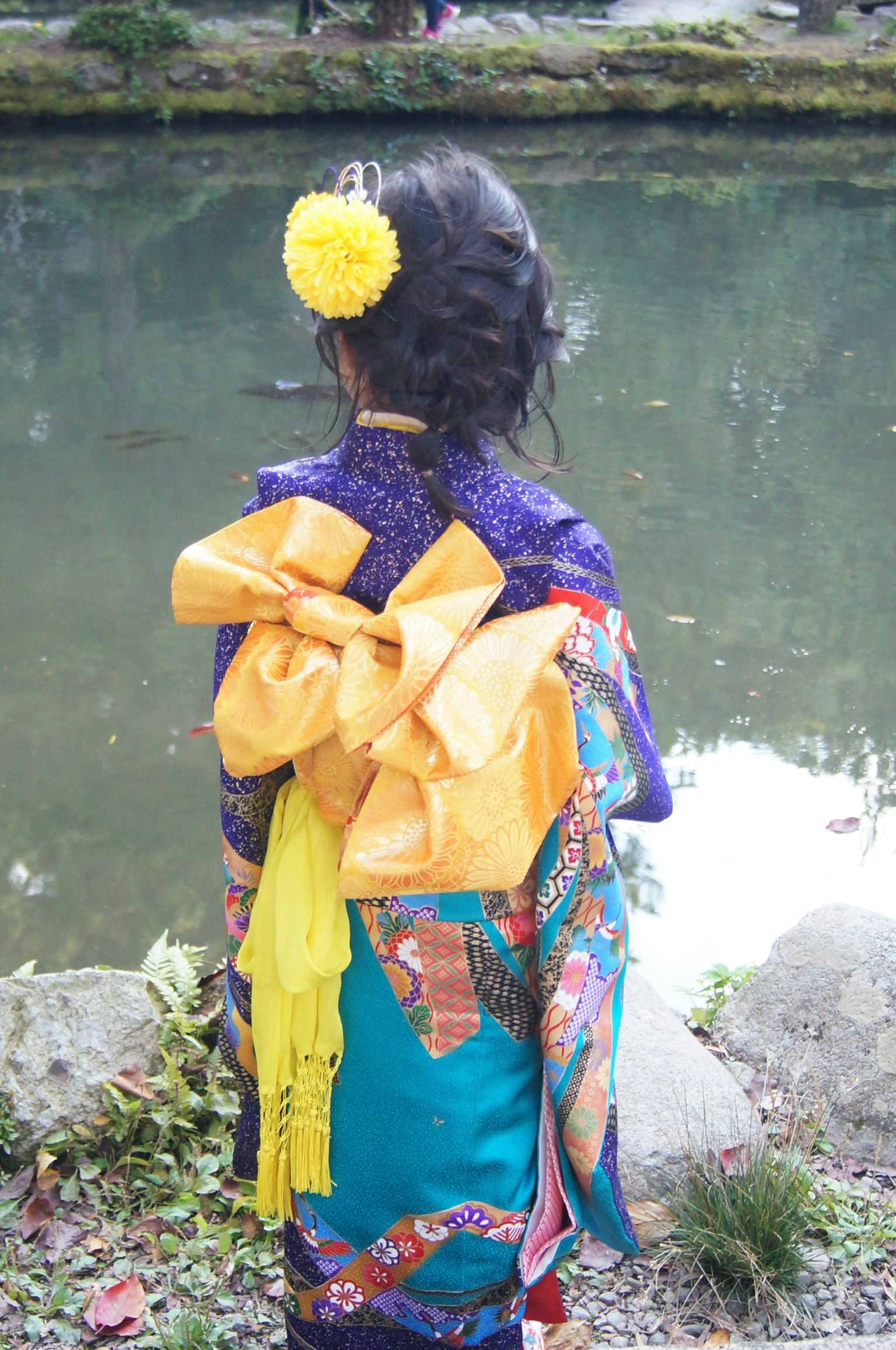 Seorang wanita terlihat dari belakang mengenakan kimono cerah dengan warna biru dan ungu serta ikatan kuning besar dan hiasan kuning di rambutnya