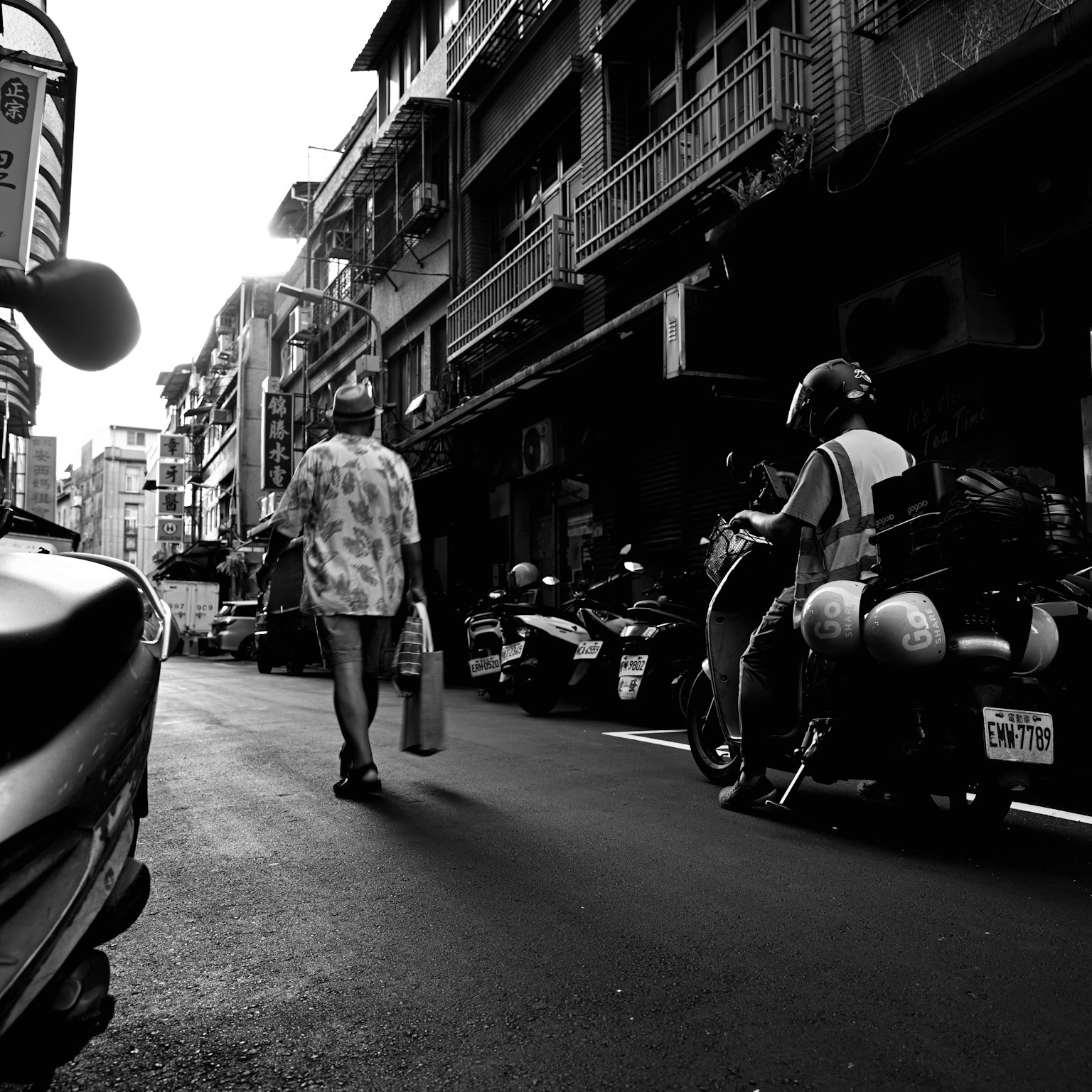 Un homme marchant dans une rue étroite avec des motos garées dans un cadre urbain en noir et blanc