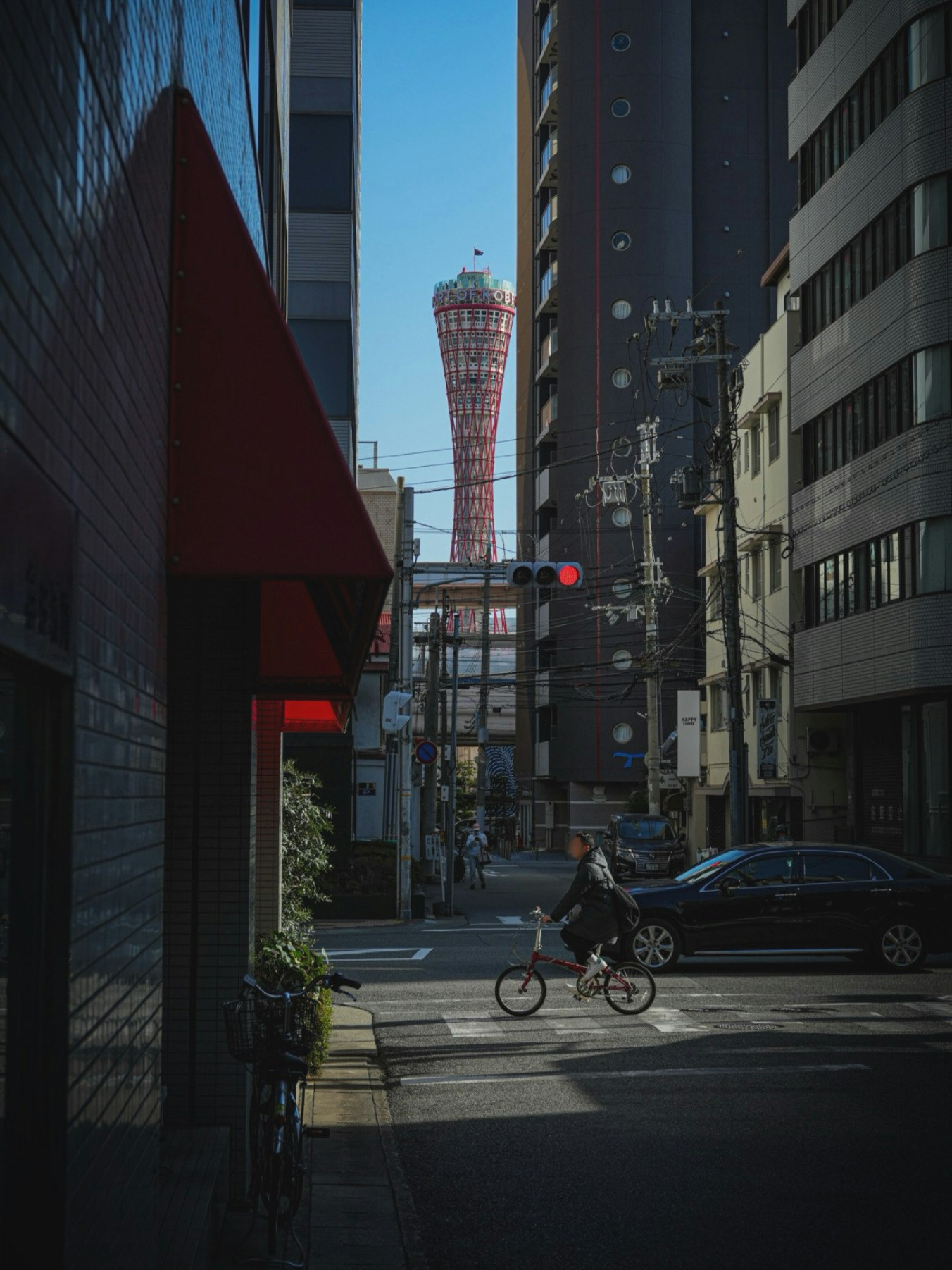 狭い通りに自転車に乗った人物と高層ビルが並ぶ風景の中に、遠くに通天閣が見える
