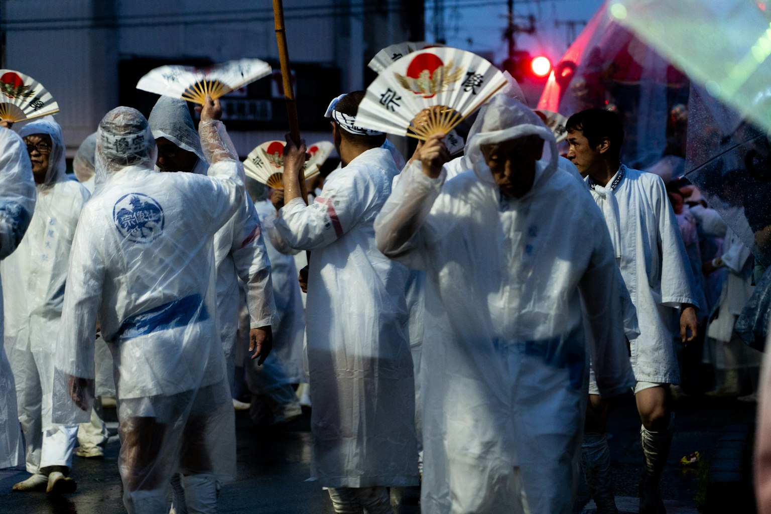 Procession of people in white raincoats holding fans in the rain