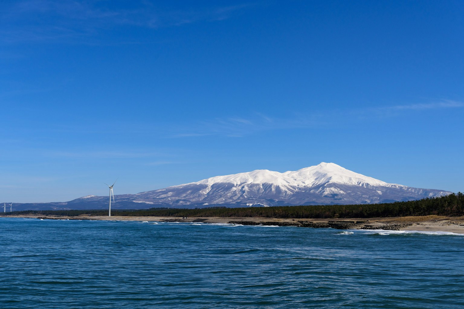 Landschaftsansicht eines schneebedeckten Berges und des Meeres unter einem blauen Himmel mit einer Windkraftanlage