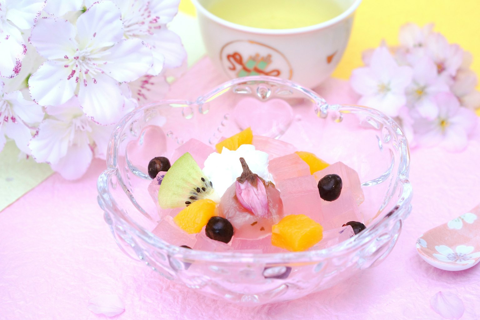 Postre colorido con frutas y gelatina en un bol transparente fondo rosa con flores de cerezo y una taza de té