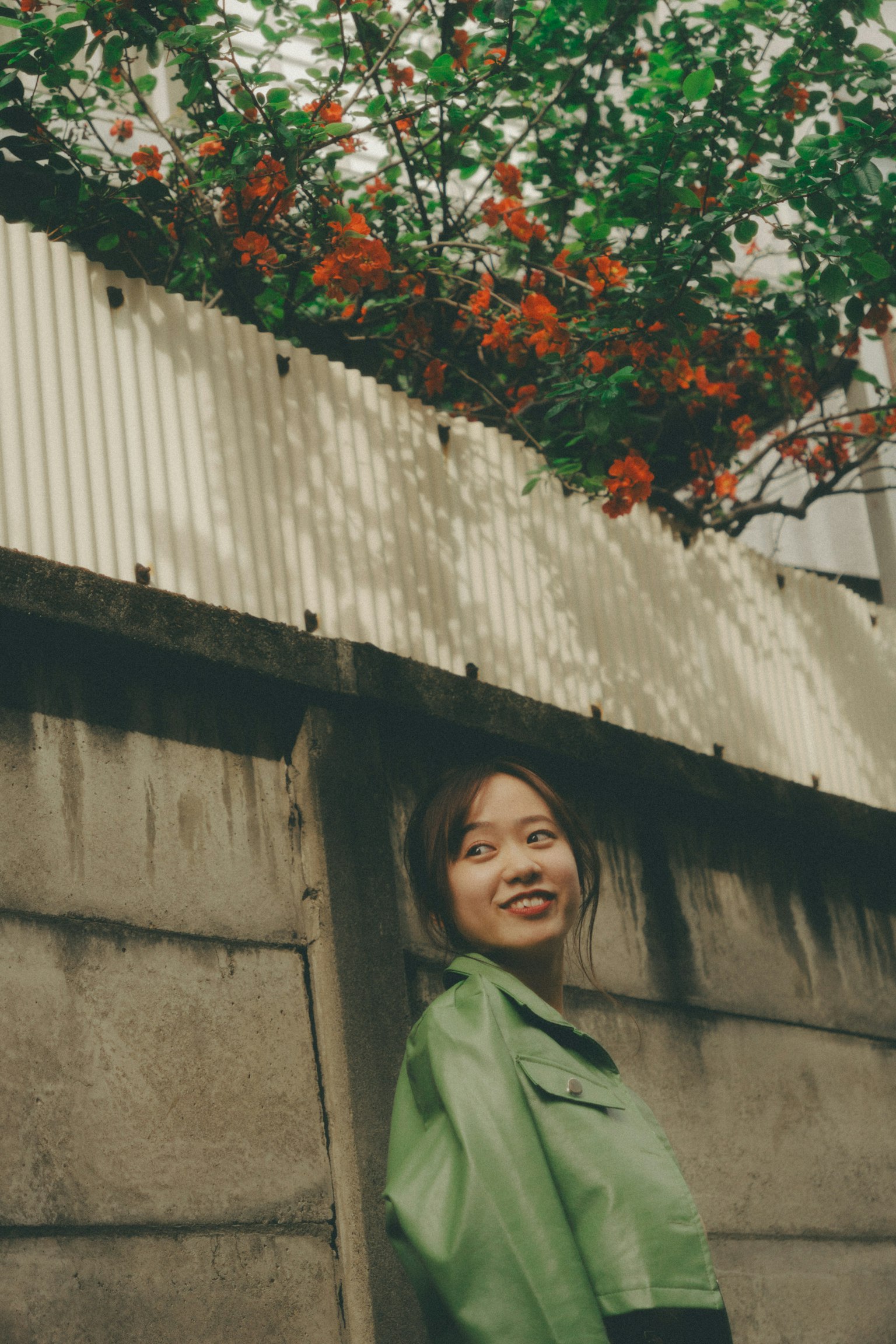 Femme en veste verte se tenant près d'un mur avec des plantes à fleurs orange au-dessus