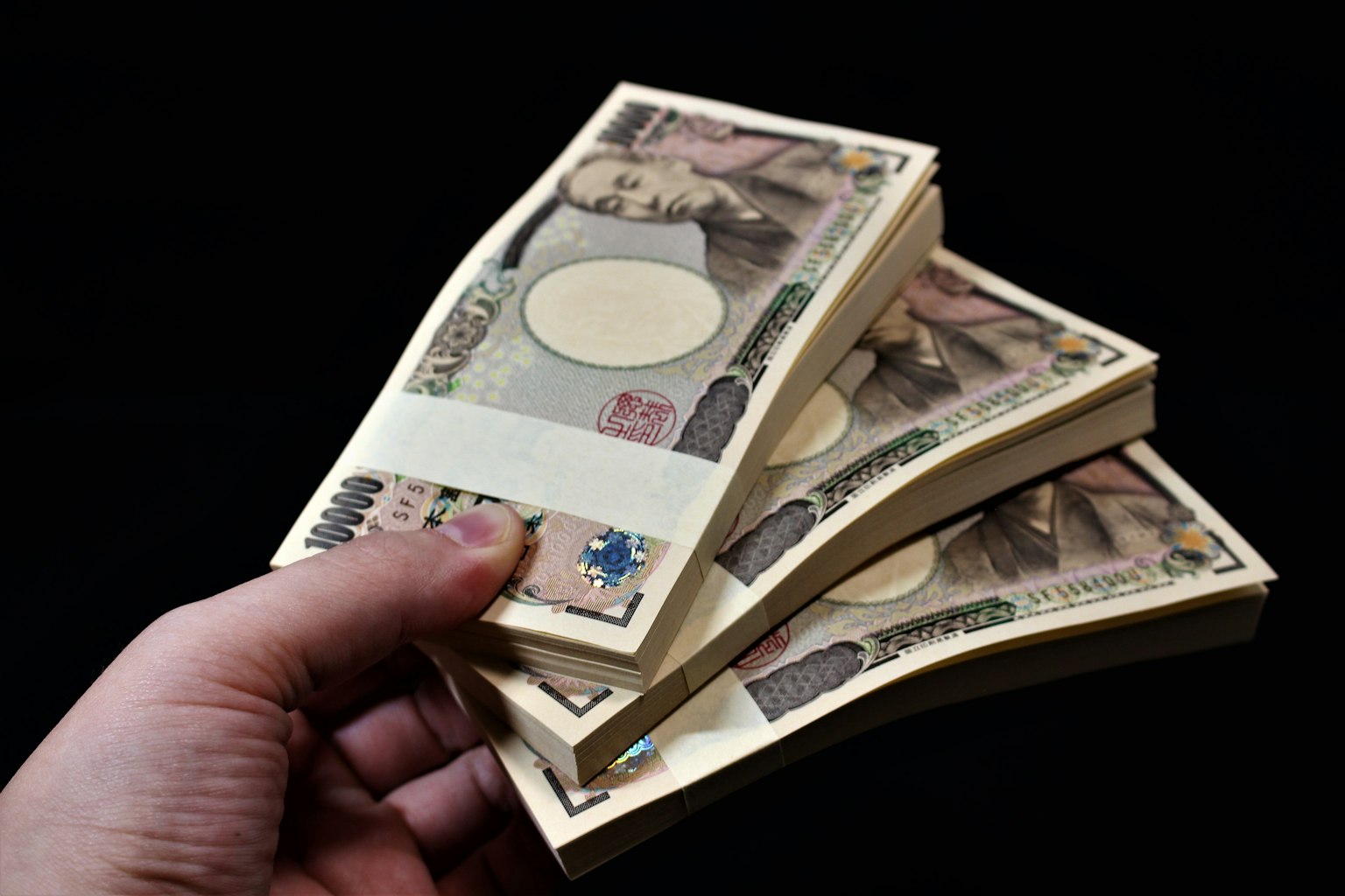 A hand holding three bundles of Japanese yen notes against a black background