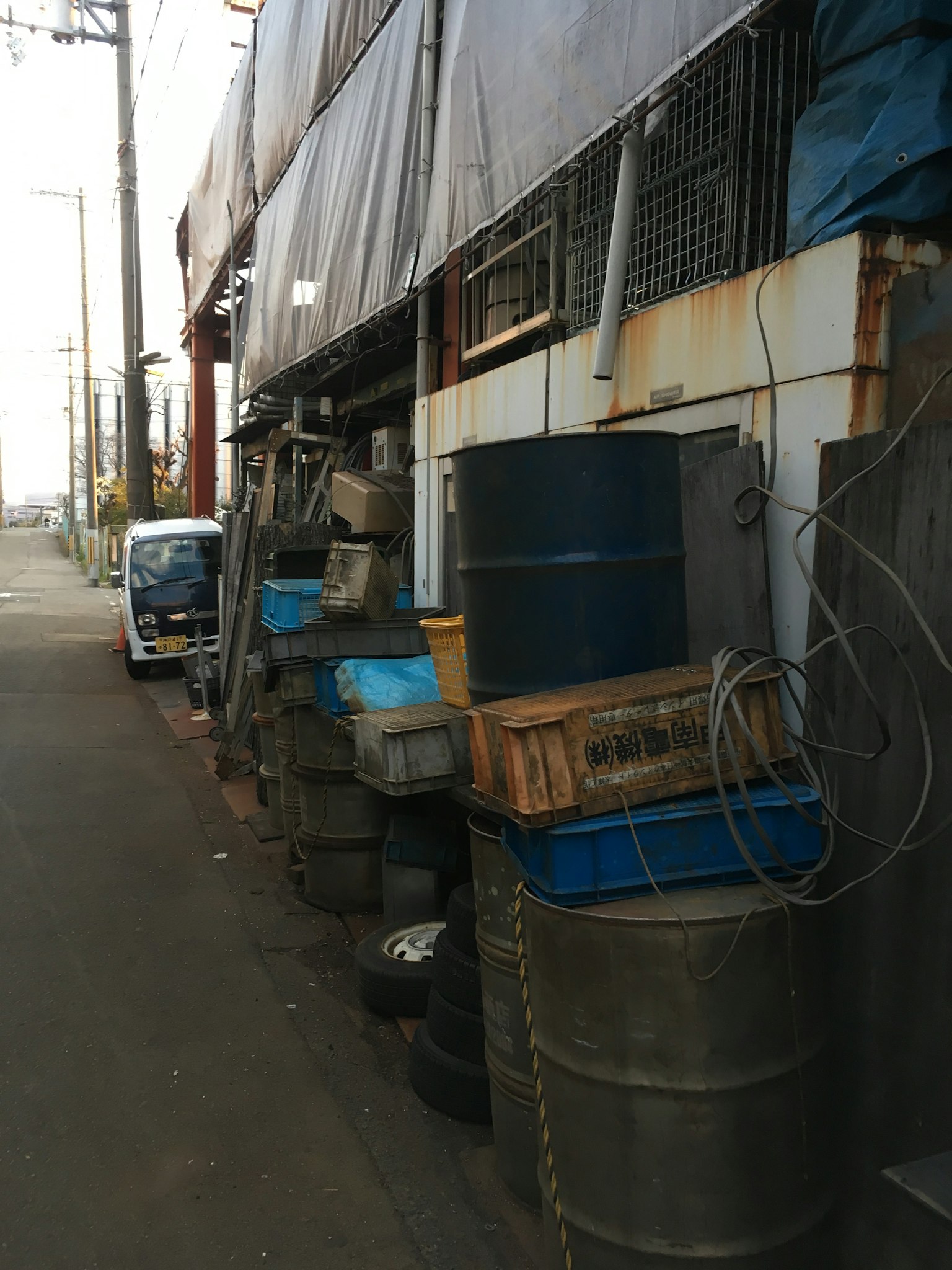 ฉากเขตอุตสาหกรรมที่มีถังสีน้ำเงินและกล่องไม้เรียงรายอยู่ตามถนนแคบ