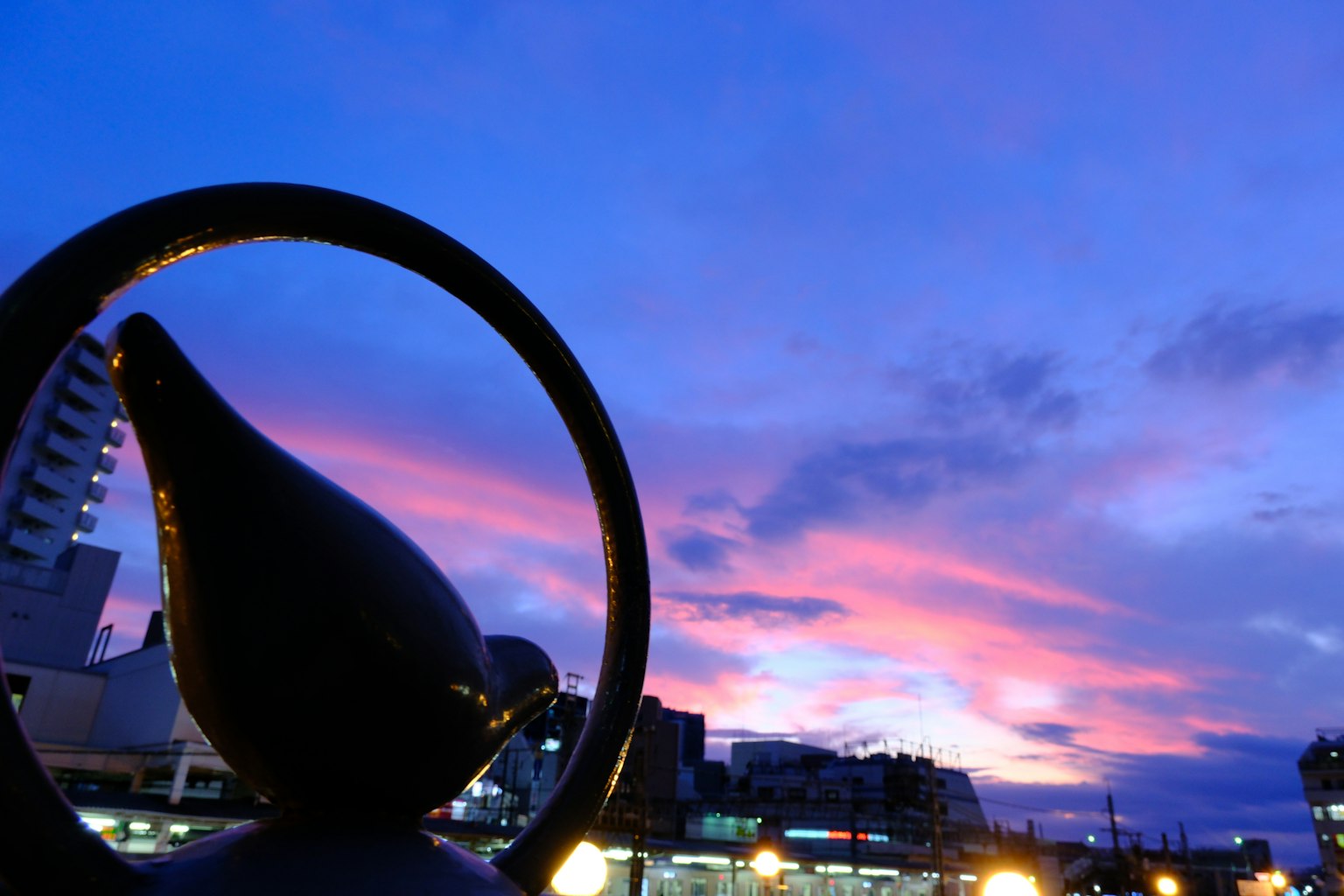 Escultura negra con diseño circular contra un cielo de atardecer colorido y un paisaje urbano