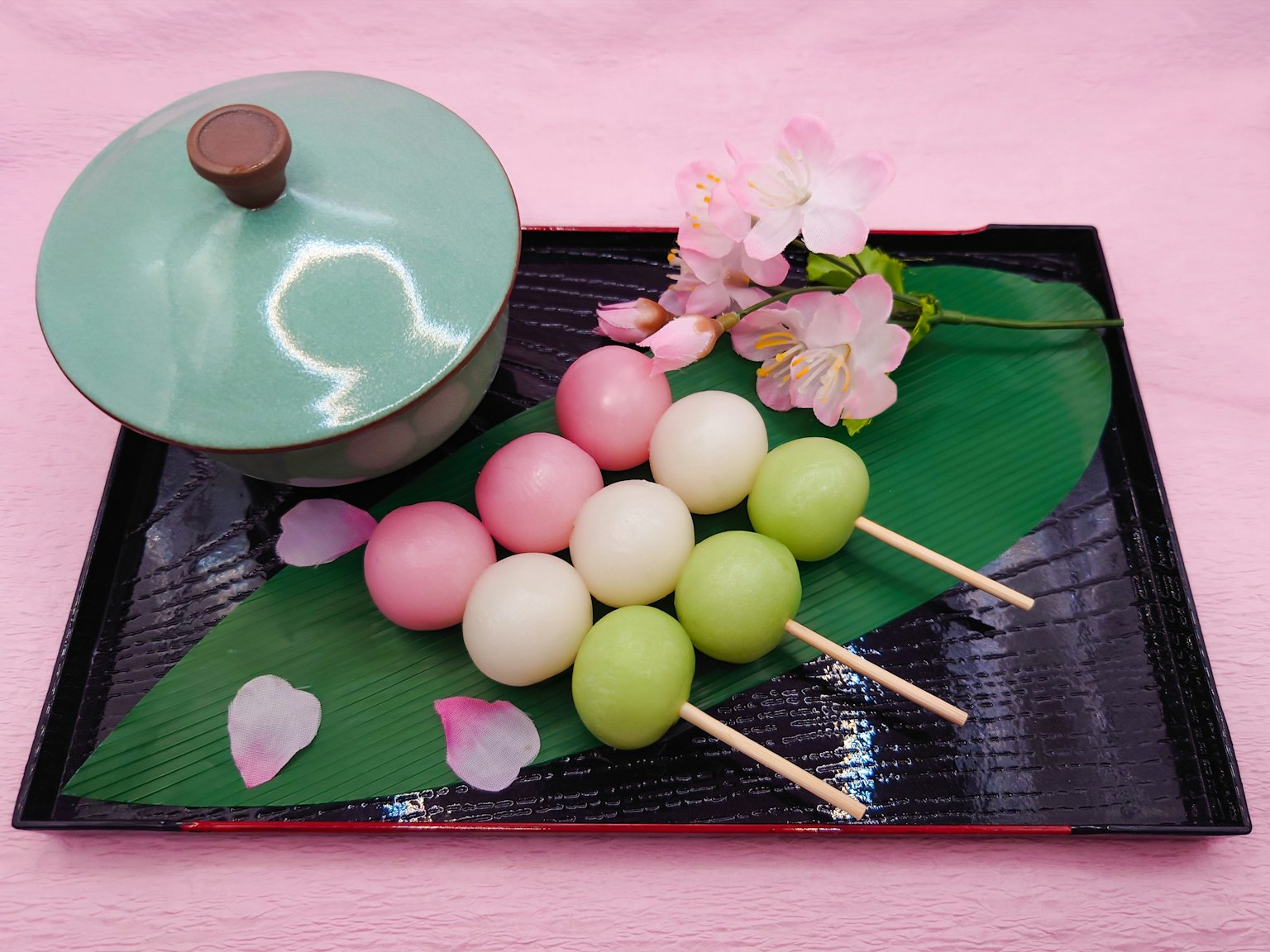 Colorful dango skewers arranged on a black tray with a green lidded bowl and cherry blossoms
