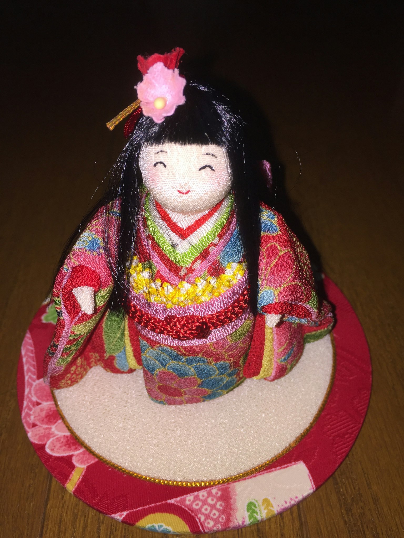 Một con búp bê truyền thống của Nhật Bản mặc kimono đầy màu sắc ngồi trên một đế tròn màu đỏ