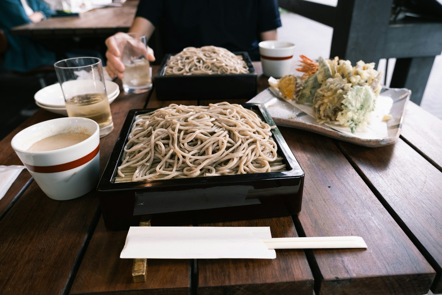 桌子上摆放着荞麦面和天妇罗菜肴，背景中有饮料和其他食物