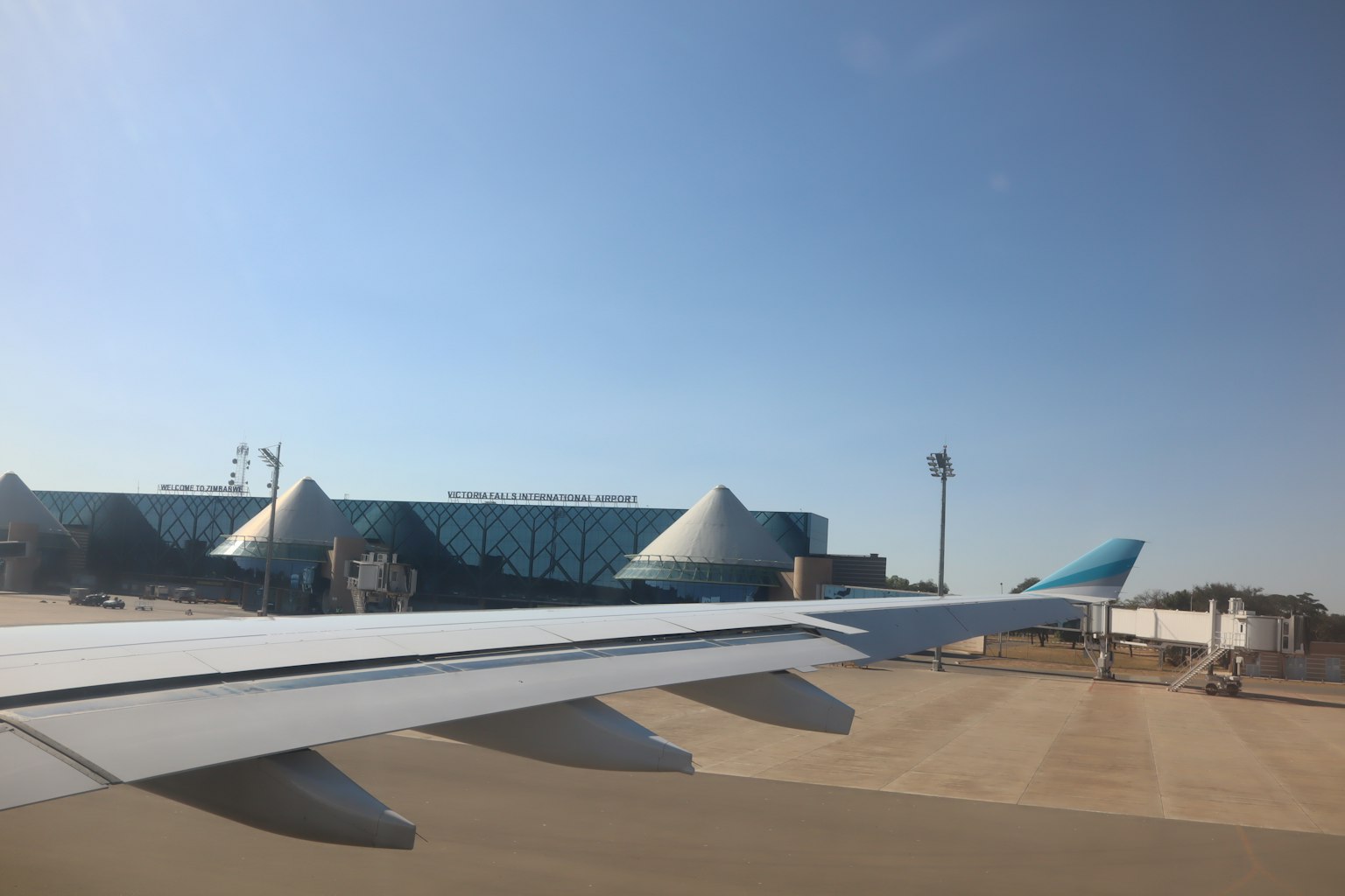 Vista dell'ala di un aereo con un edificio aeroportuale sullo sfondo e cielo blu chiaro