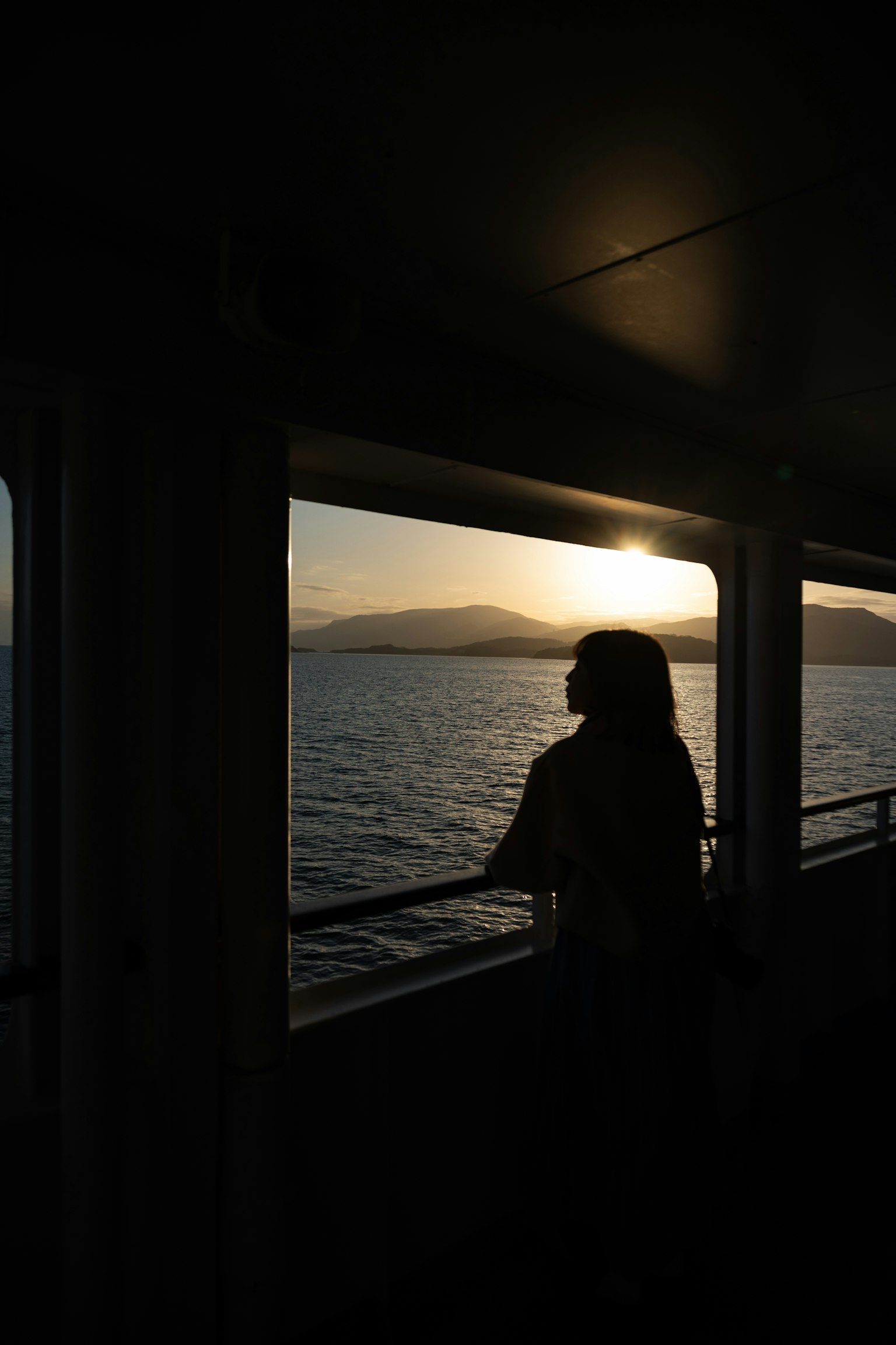Siluet seorang wanita melihat matahari terbenam dari jendela kapal dengan pemandangan laut dan gunung