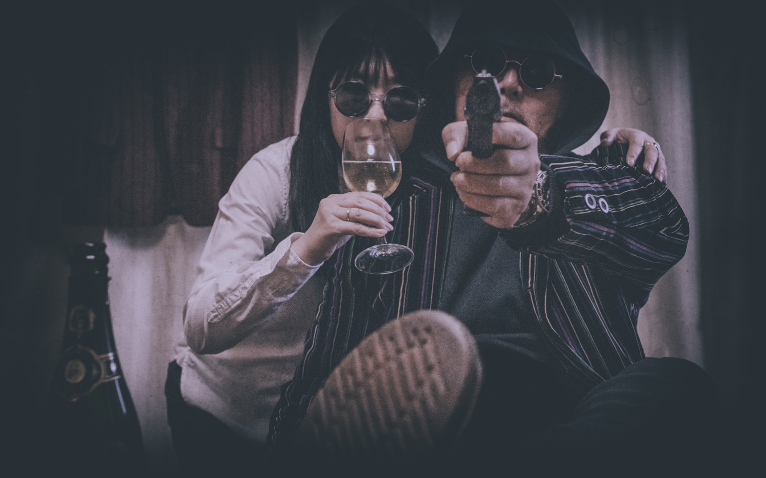 A dark-themed image featuring a man and woman wearing sunglasses with the woman holding a wine glass and the man aiming a gun at the camera