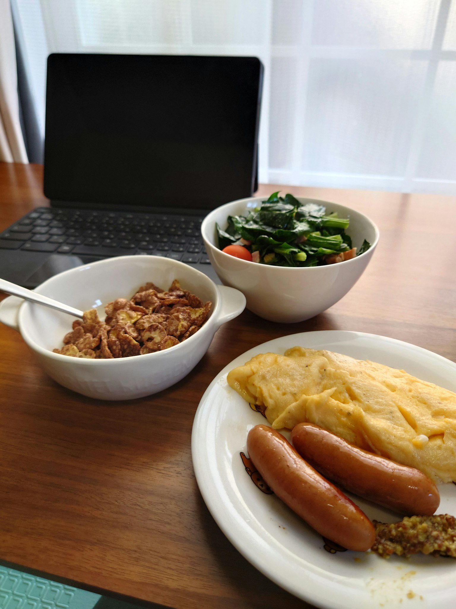Plato de desayuno con tortilla salchichas y ensalada con una computadora portátil al fondo