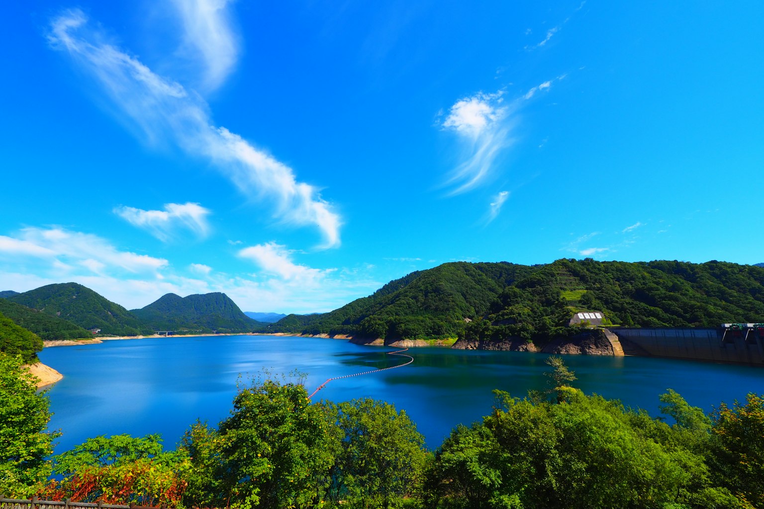 Vista escénica de un lago de represa rodeado de montañas verdes y un cielo azul claro
