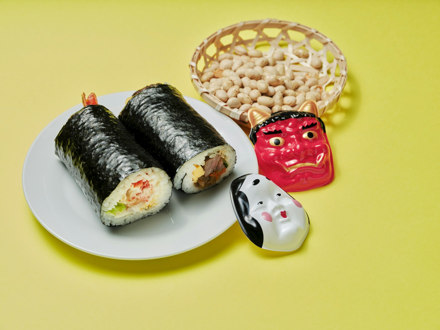 Gulungan sushi di piring putih dengan keranjang kacang topeng iblis merah dan patung kucing putih di latar belakang kuning