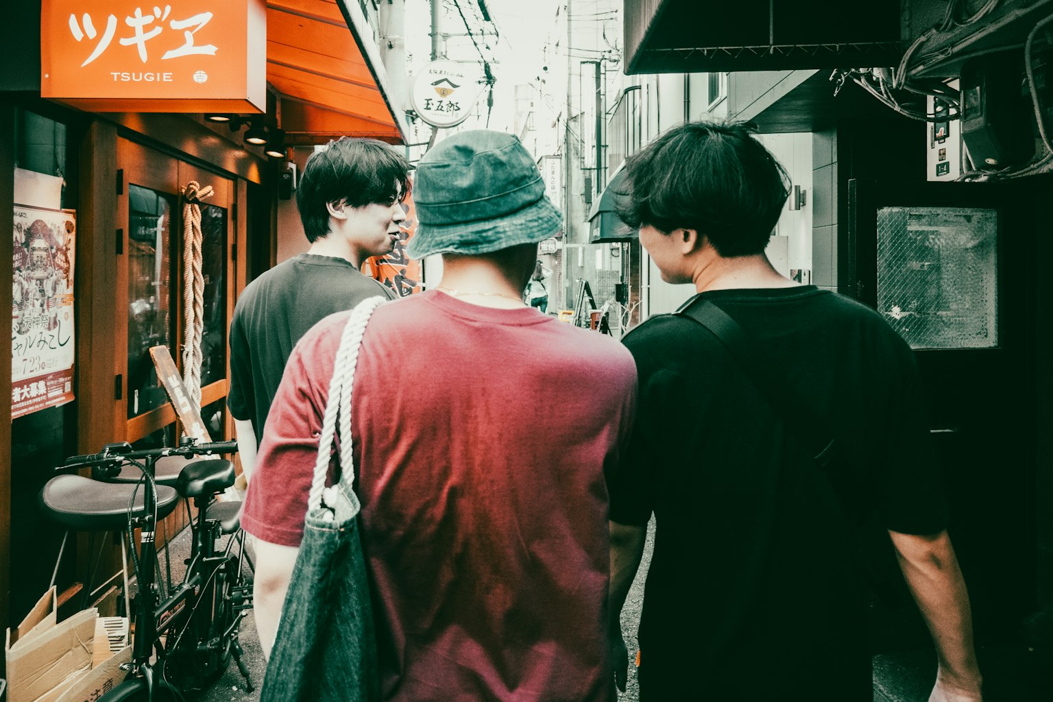 Drei junge Menschen, die eine enge Straße entlanggehen, mit Geschäftsschildern und einem Fahrrad im Hintergrund