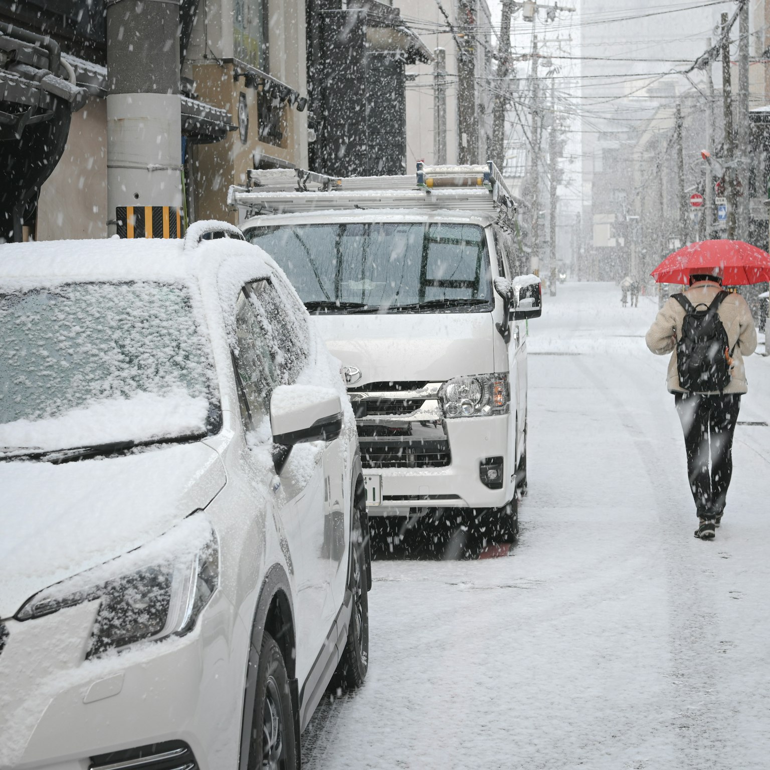 A person walking with a red umbrella in a snowy street with white cars lining the road