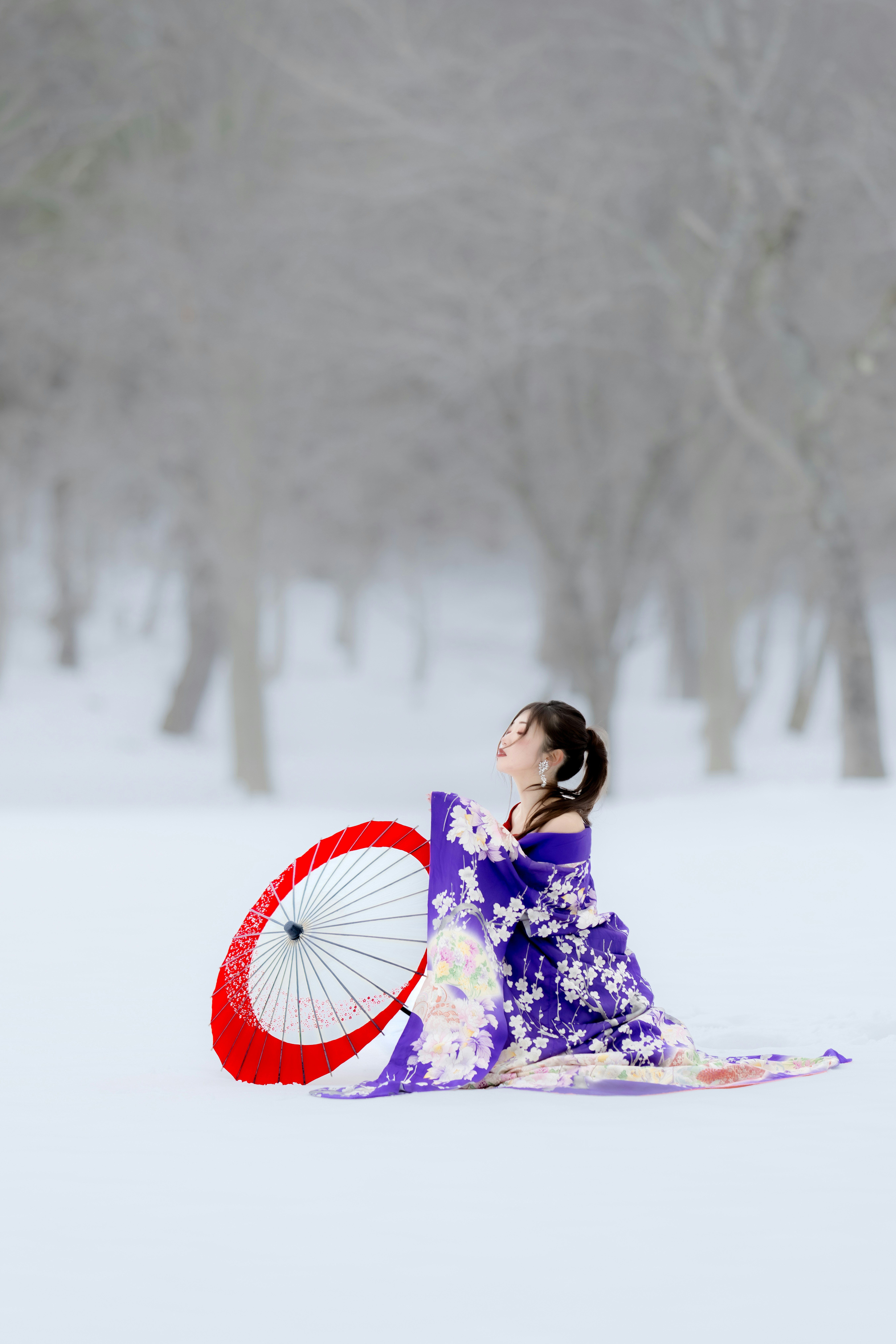 Frau in traditioneller japanischer Kleidung sitzt im Schnee mit einem Regenschirm
