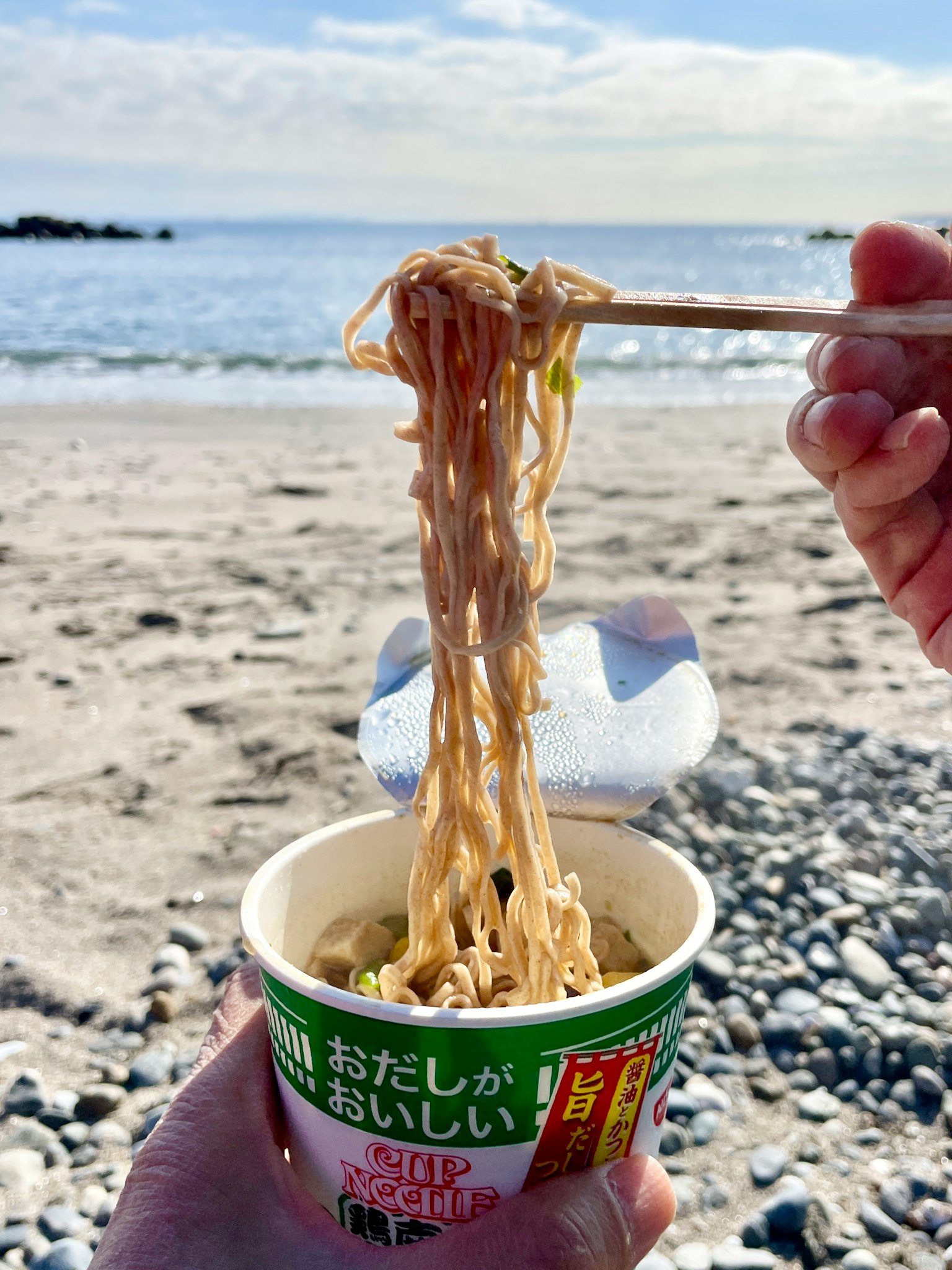 Una mano che tiene noodles da un bicchiere sulla spiaggia con sfondo marino