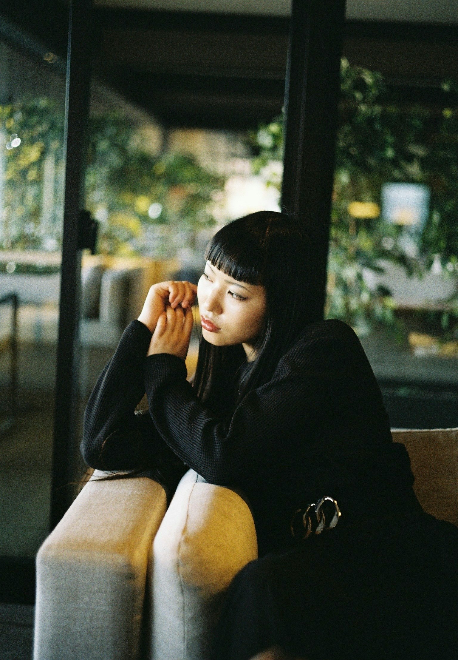 A woman sitting on a sofa looking contemplative with greenery in the background