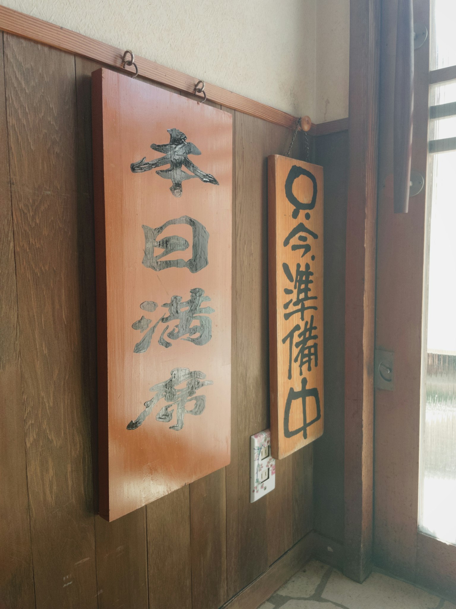 Zwei Schilder hängen an einer Holzwand Ein Schild zeigt japanische Zeichen während das andere einen hellen orangefarbenen Hintergrund mit weißem Text hat
