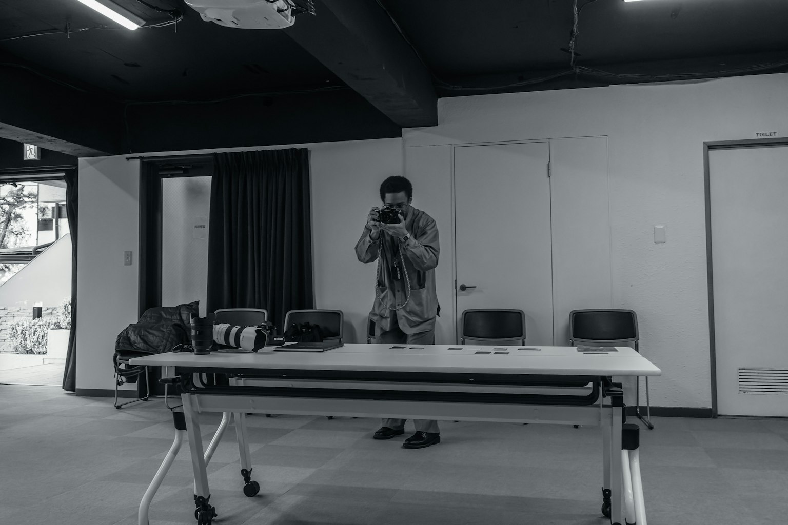 黑白圖像中，一個男人在房間裡站著，手裡拿著相機 桌子上放著相機和包