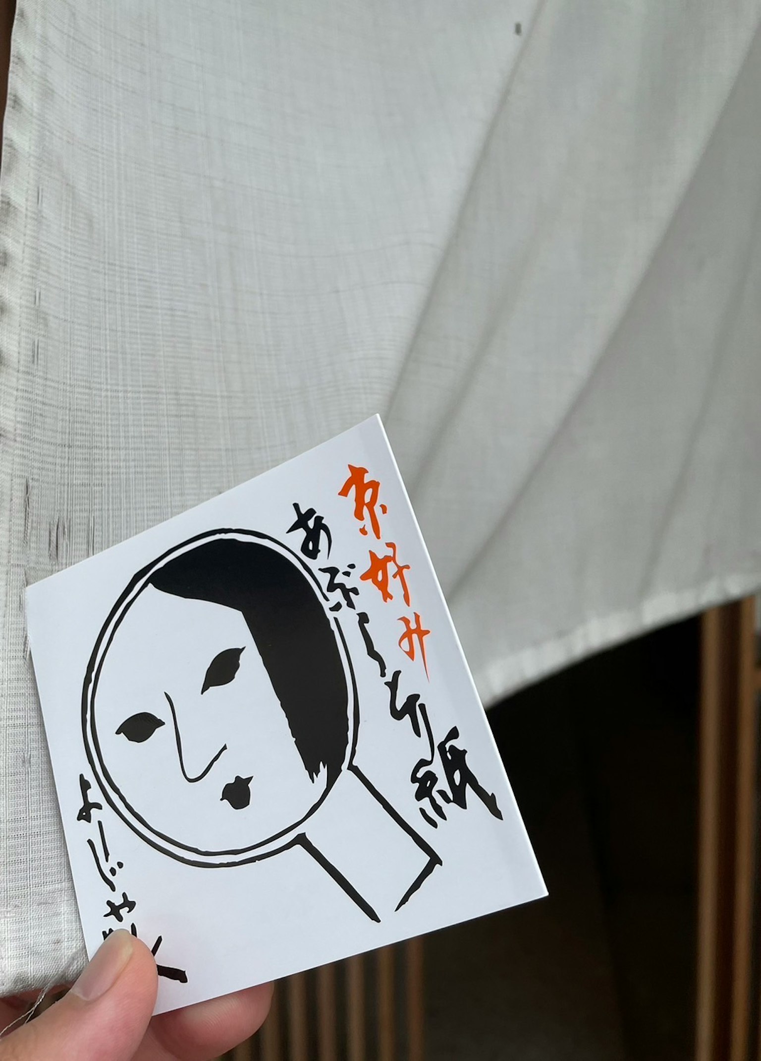 一隻手在白色布料前拿著插圖卡片，顯著的面部設計和日文文本