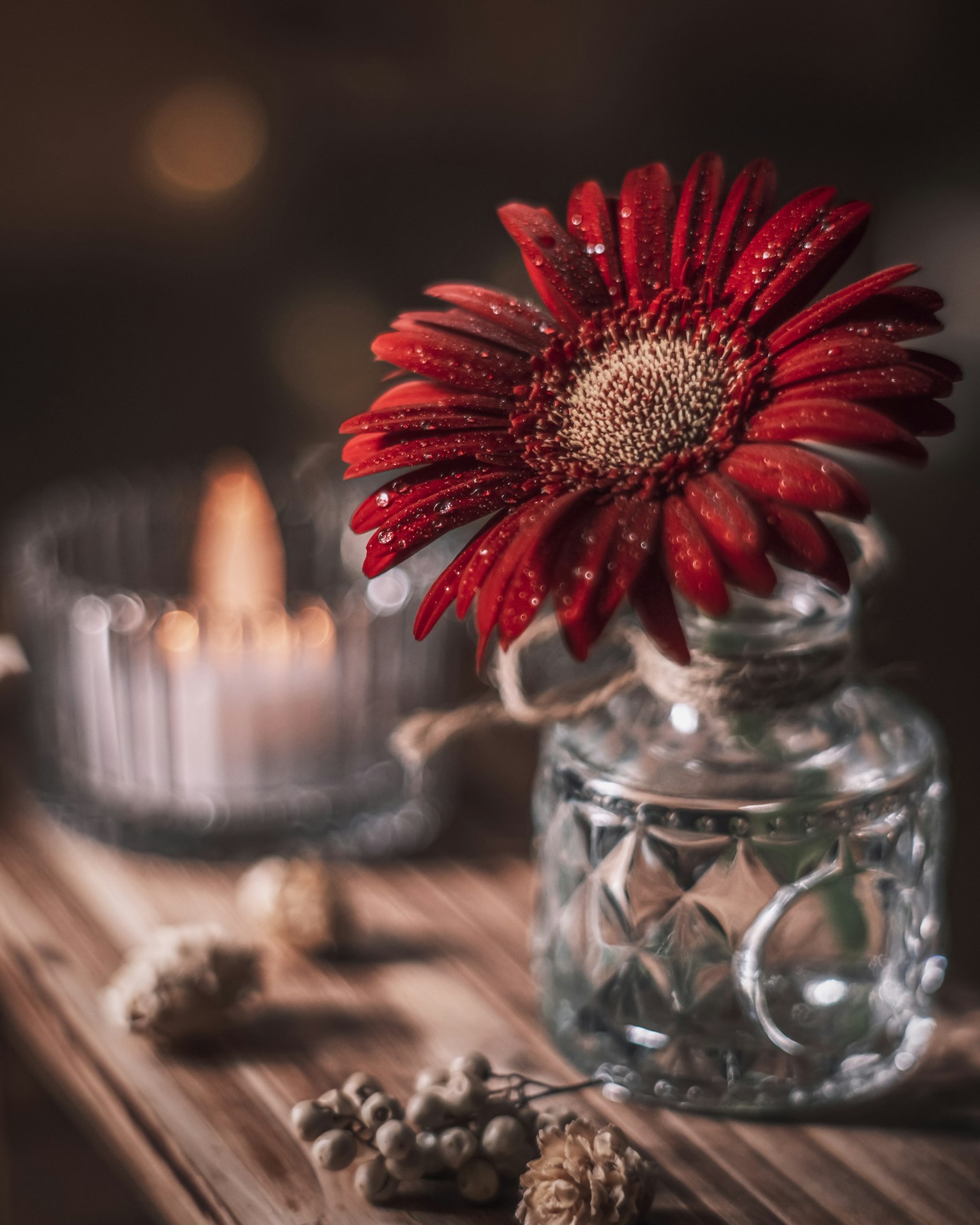 A red flower in a clear glass vase with a candle on a wooden table