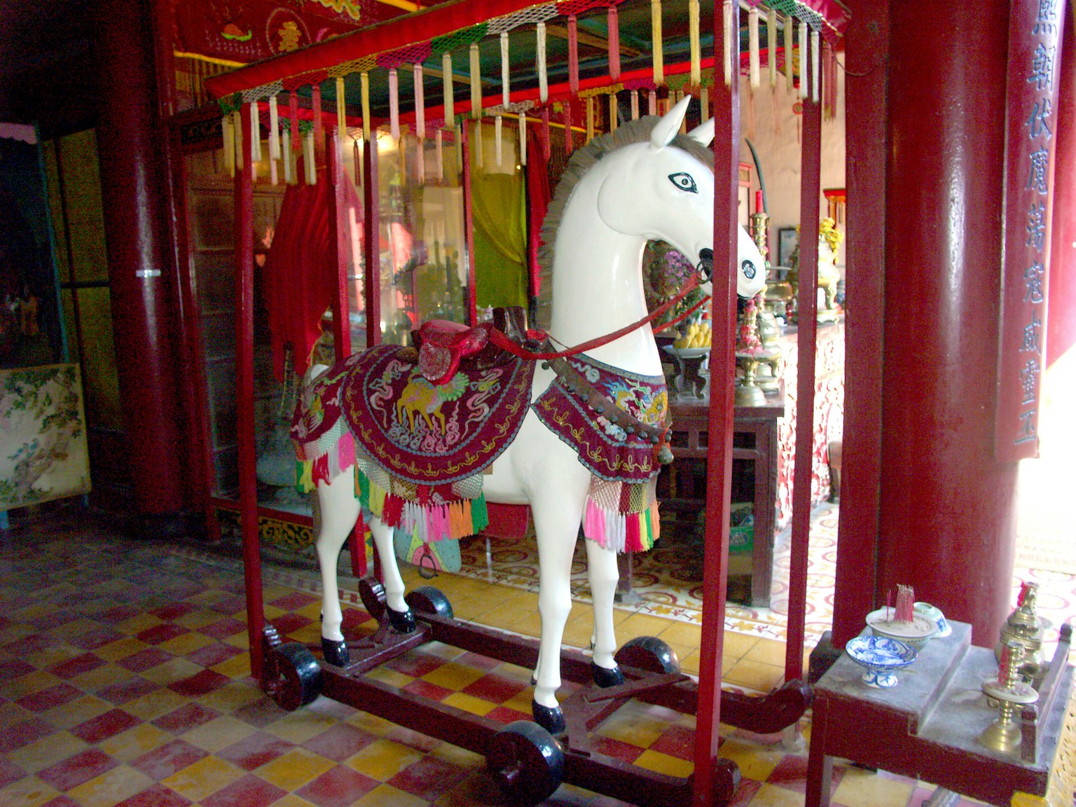 Escena interior con una estatua de caballo de madera blanca adornada con una silla colorida y elementos decorativos contra pilares rojos