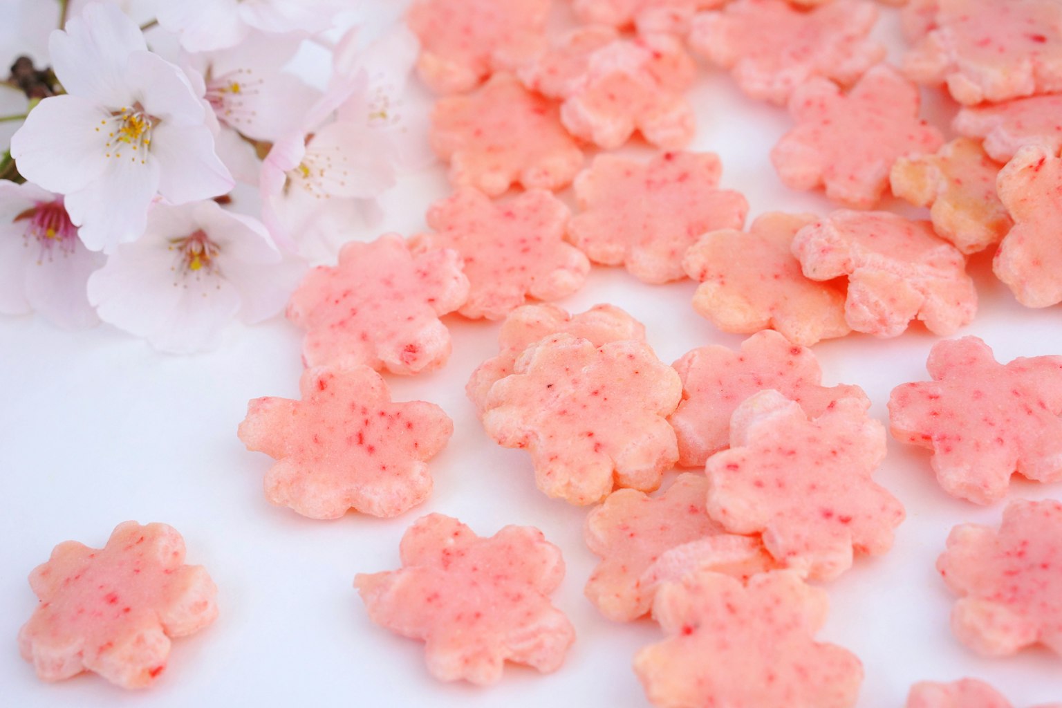 Dolcetti rosa a forma di fiore sparsi su uno sfondo bianco con petali di ciliegio