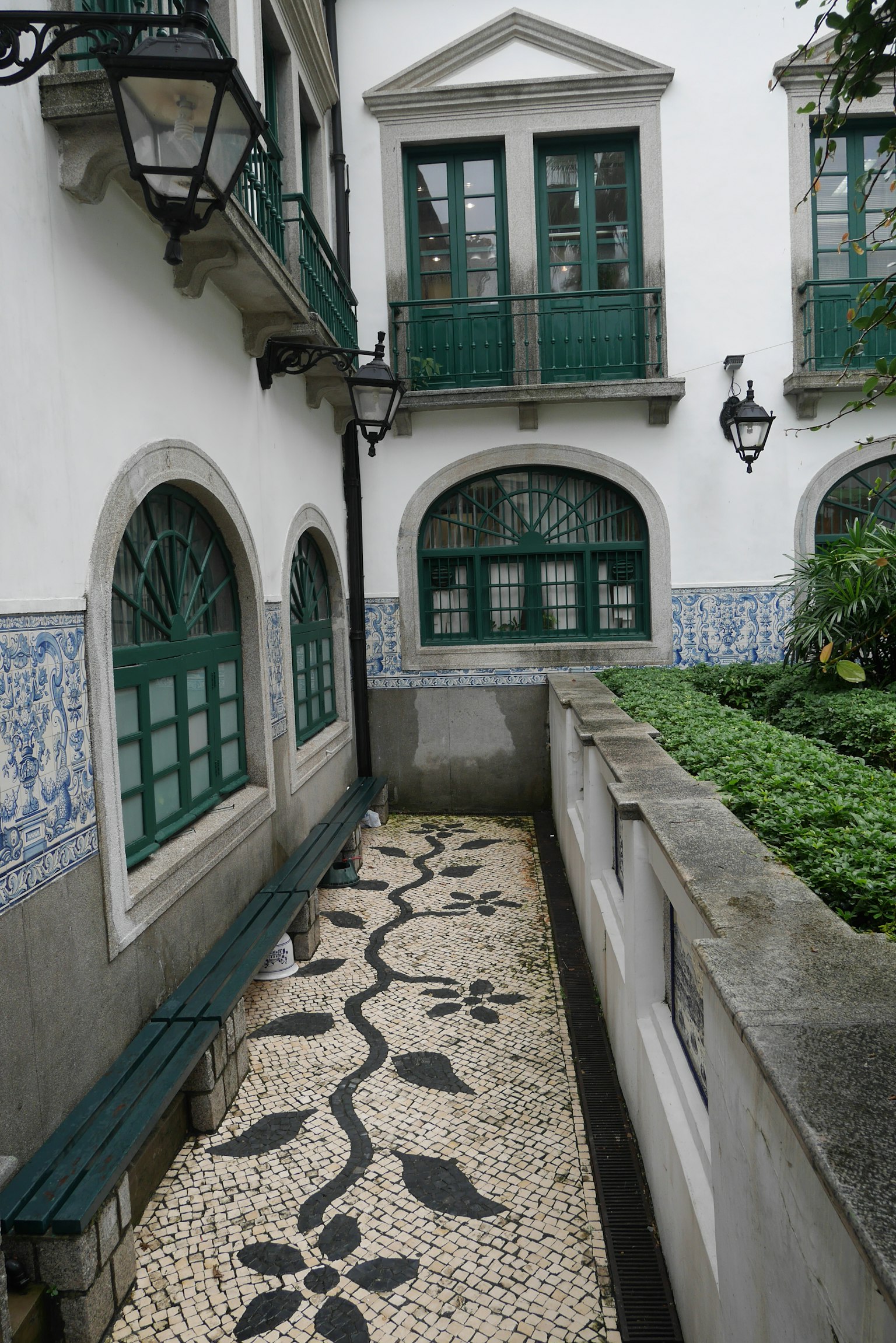Sentier étroit avec mosaïque florale entre un bâtiment blanc et une structure à fenêtres vertes