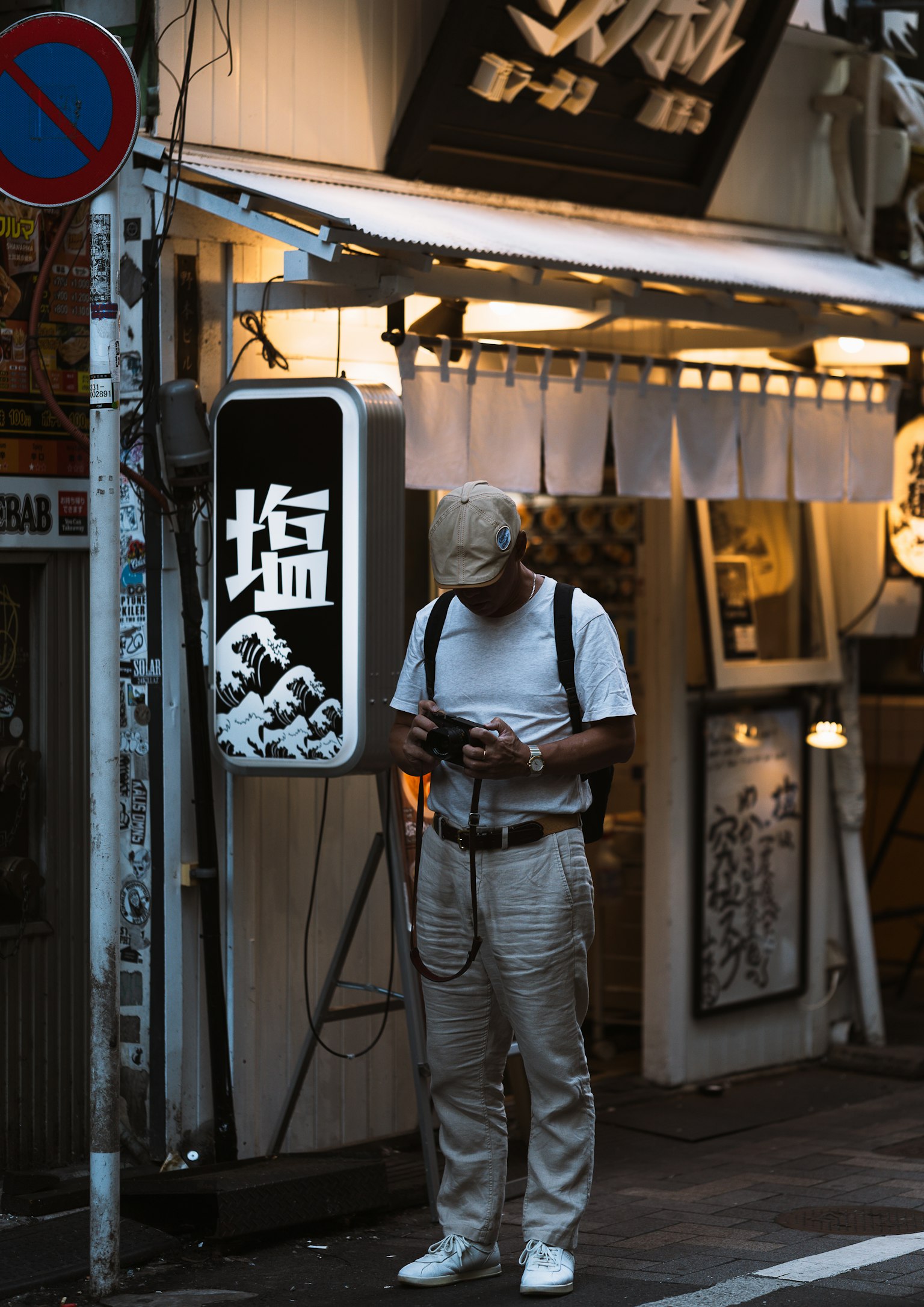 一个人在昏暗的街角看着手机，穿着白色T恤和裤子，附近有日本餐厅的招牌