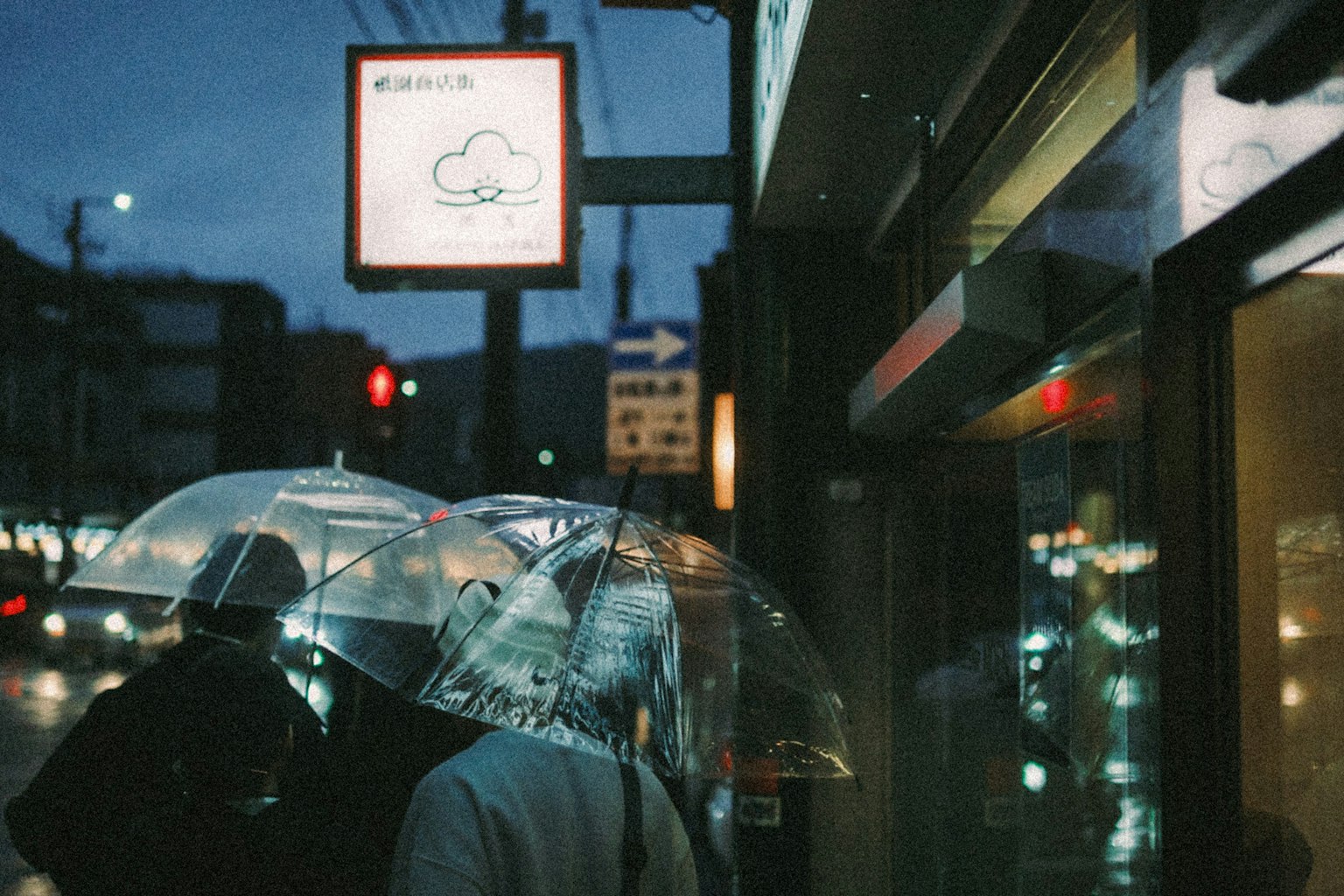 Des gens marchant avec des parapluies transparents dans une ambiance nocturne urbaine avec un panneau lumineux