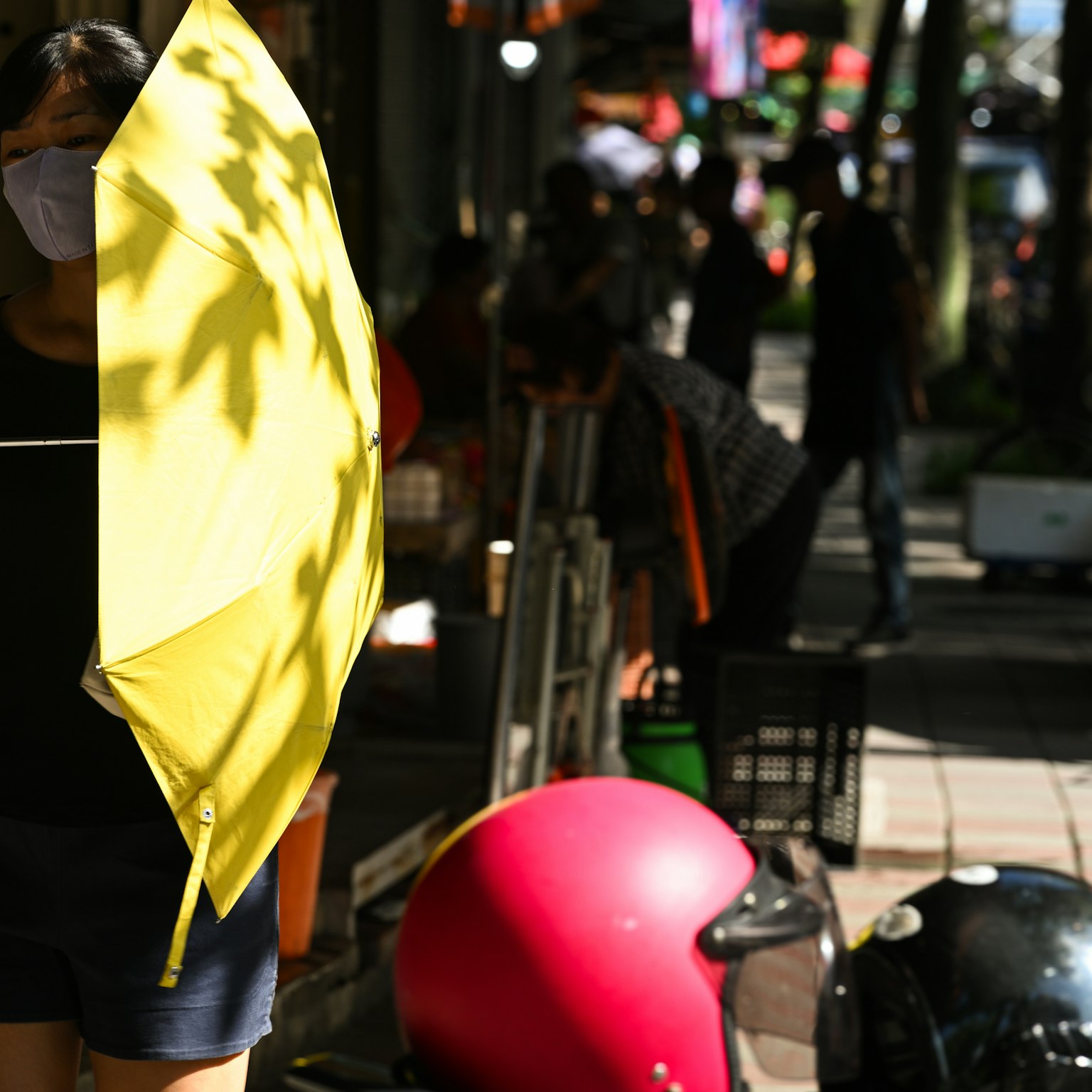 A woman holding a yellow umbrella stands in a bustling street scene with people and various objects around