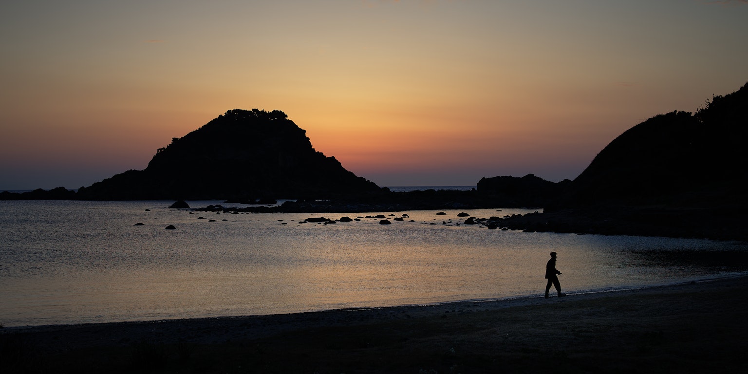 เงาคนเดินอยู่ริมชายหาดในยามพระอาทิตย์ตกกับภูเขาในพื้นหลัง