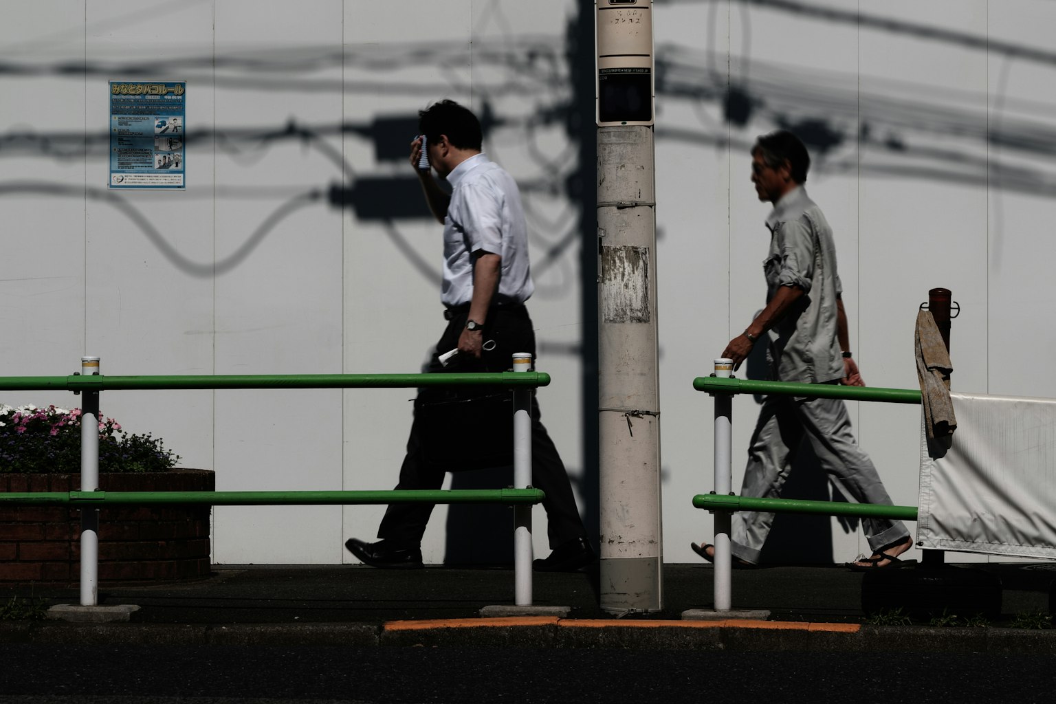 两名男性在阴影中行走，背景是白色墙壁和电线