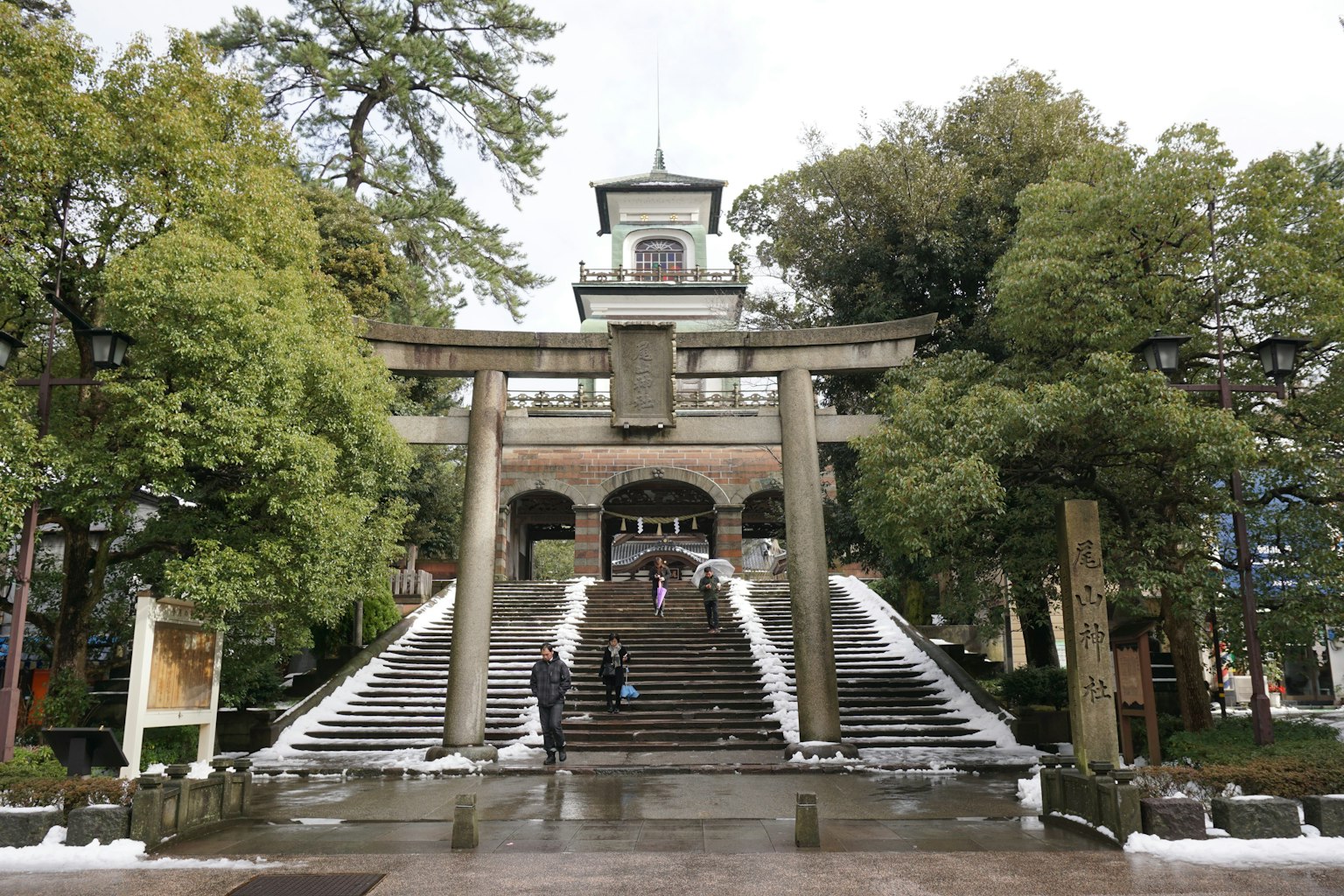 一個神社的風景，有一個大型鳥居和階梯，周圍是綠色樹木和歷史建築
