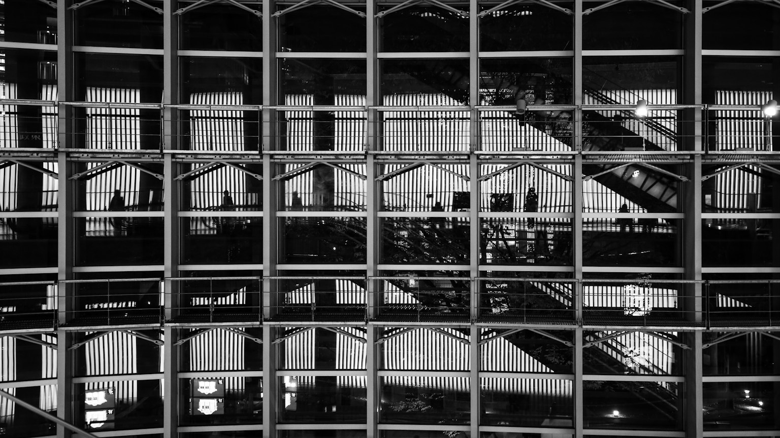 Image en noir et blanc montrant la structure intérieure d'un bâtiment moderne avec des cadres en grille verticale et des fenêtres réfléchissantes
