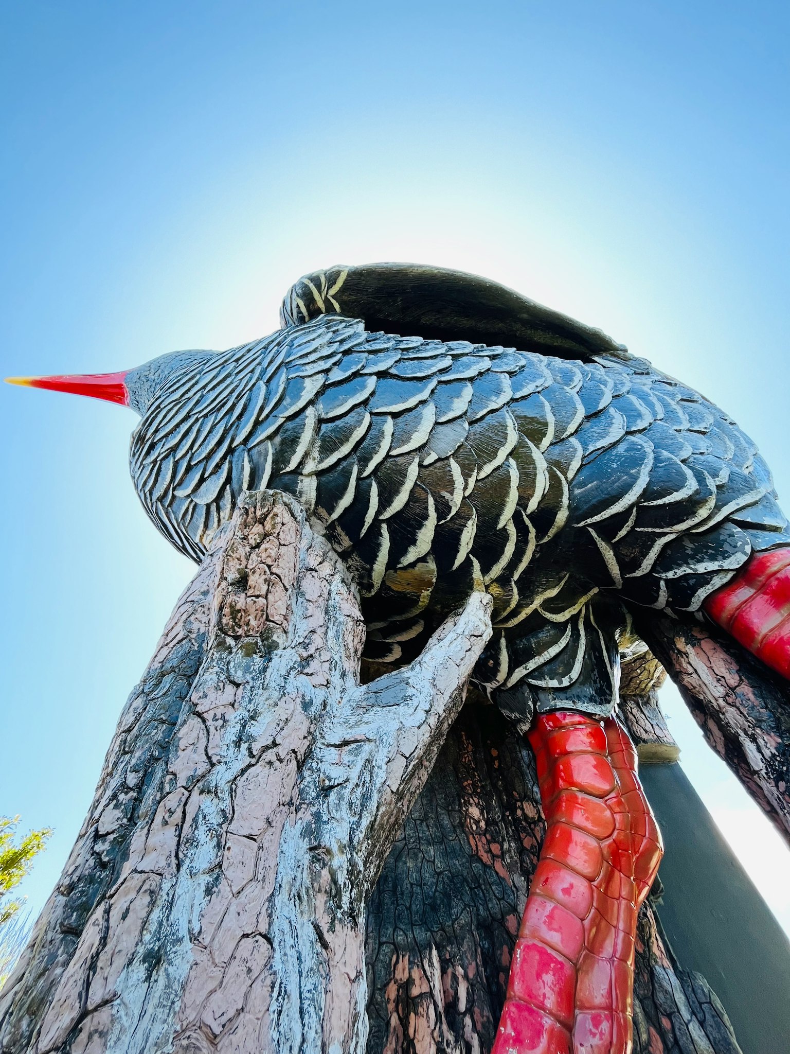 Large bird sculpture perched on a tree trunk with a prominent red beak and detailed feathers