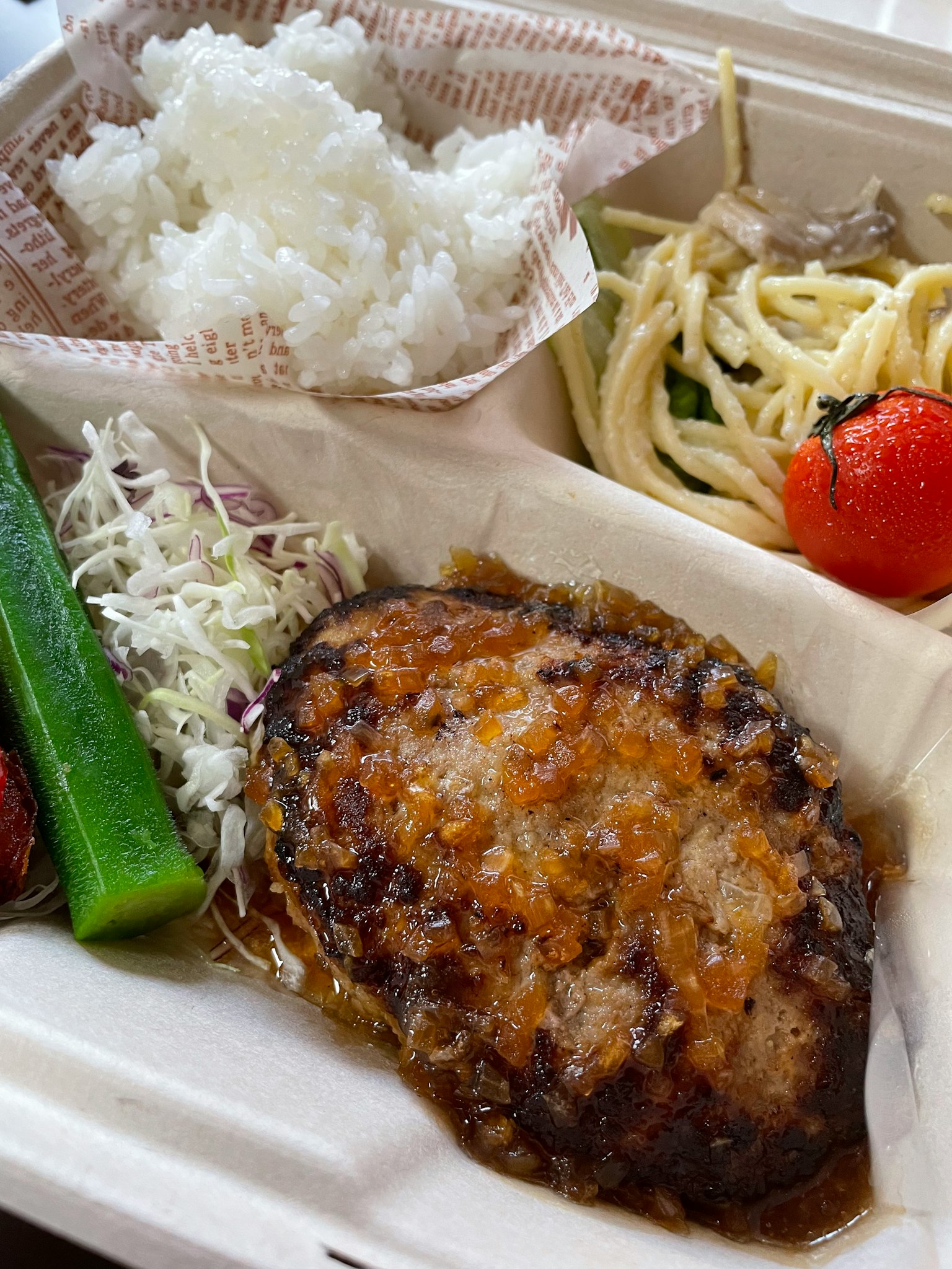 Une boîte à bento avec du riz, un steak haché, une salade, des spaghetti et une tomate