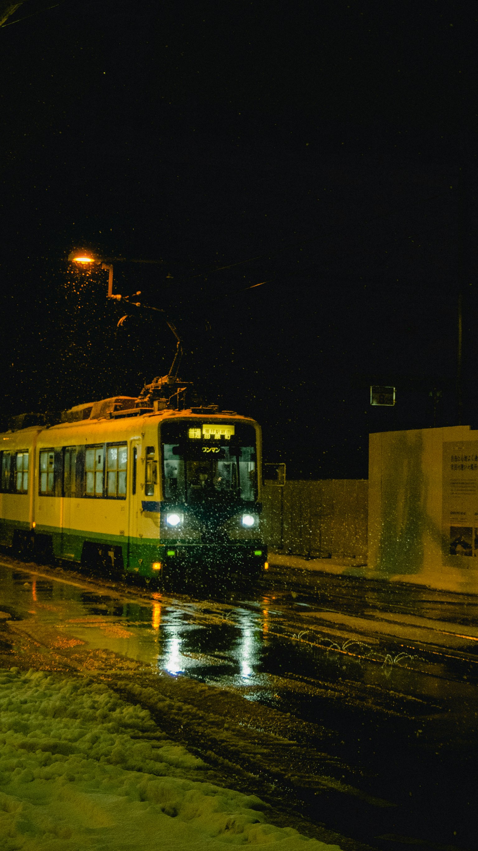 Tram hijau dan putih melintasi kota di malam hari dengan pantulan cahaya